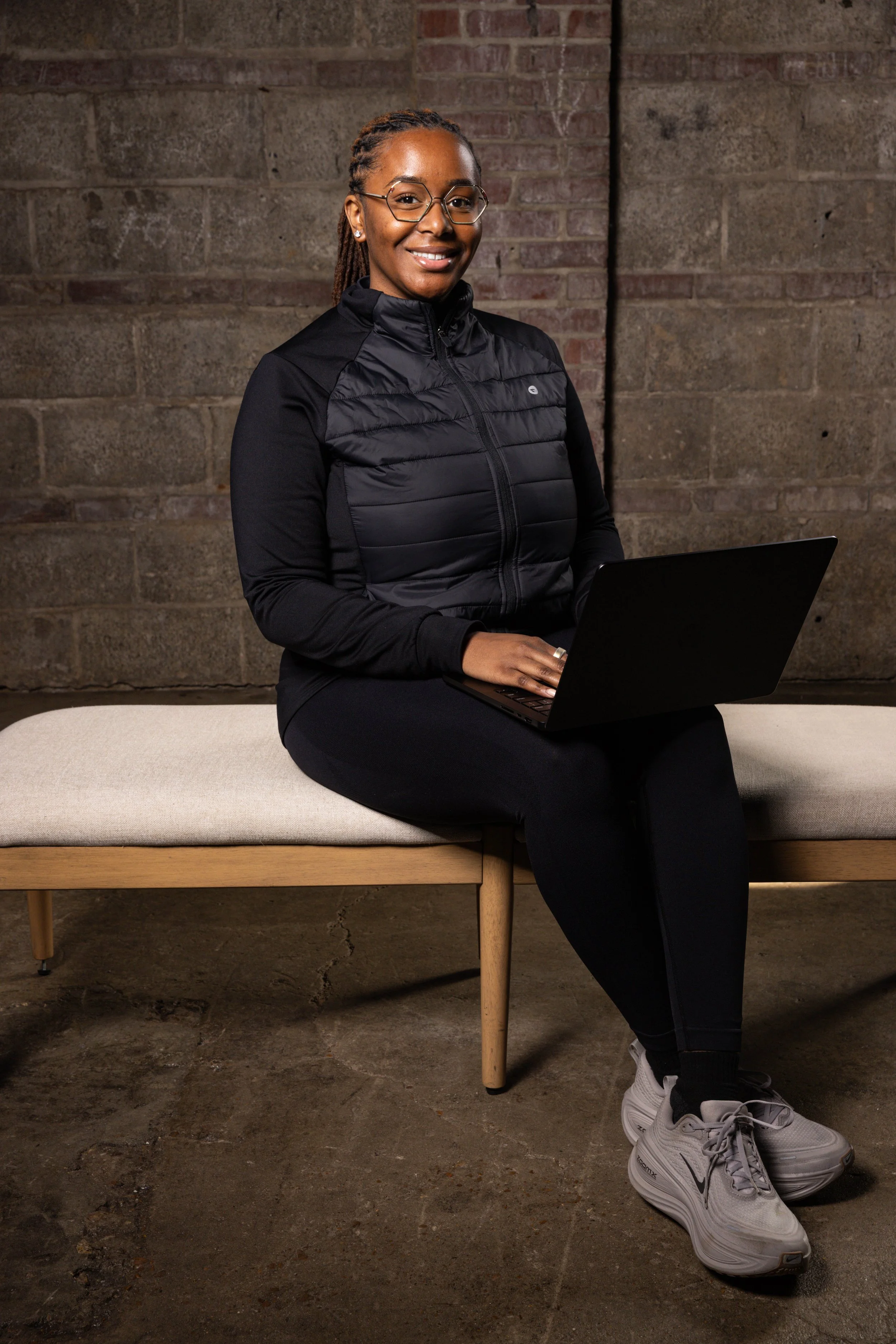 A woman sitting on a bench with a laptop, wearing glasses, a black jacket, black pants, and white Nike sneakers, in front of a brick wall.