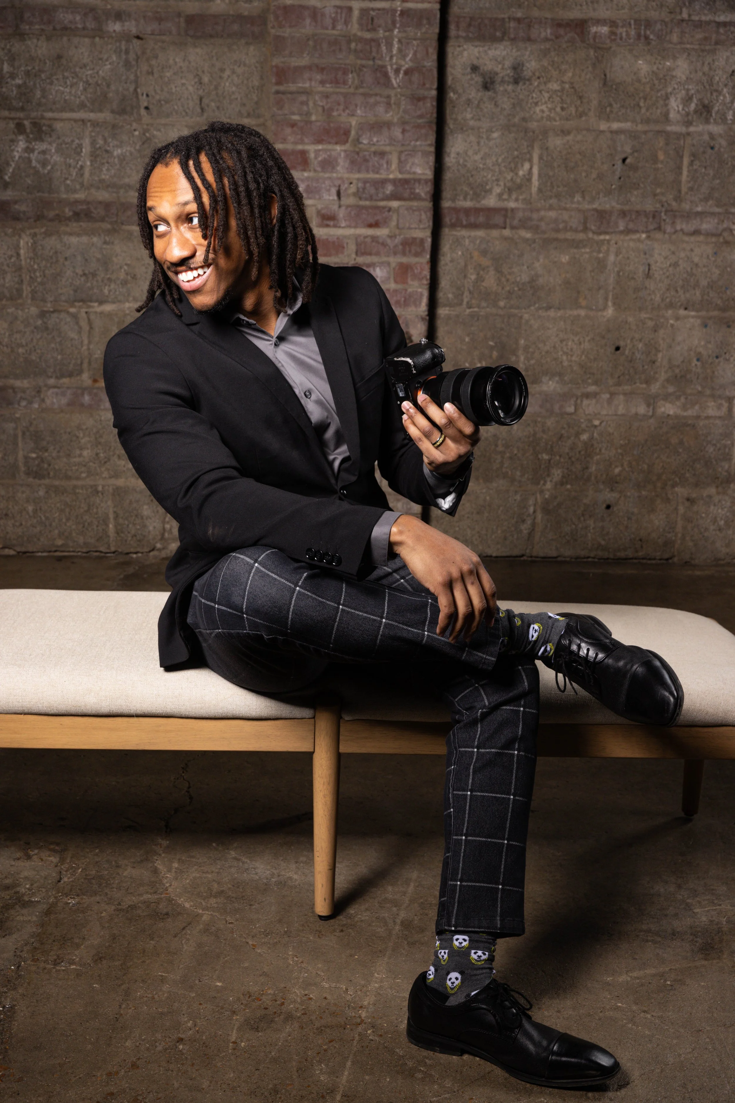 A man with dreadlocks sitting on a beige bench, holding a camera, dressed in a black blazer, gray shirt, checkered pants, black shoes, and panda-patterned socks, smiling and looking to the side.