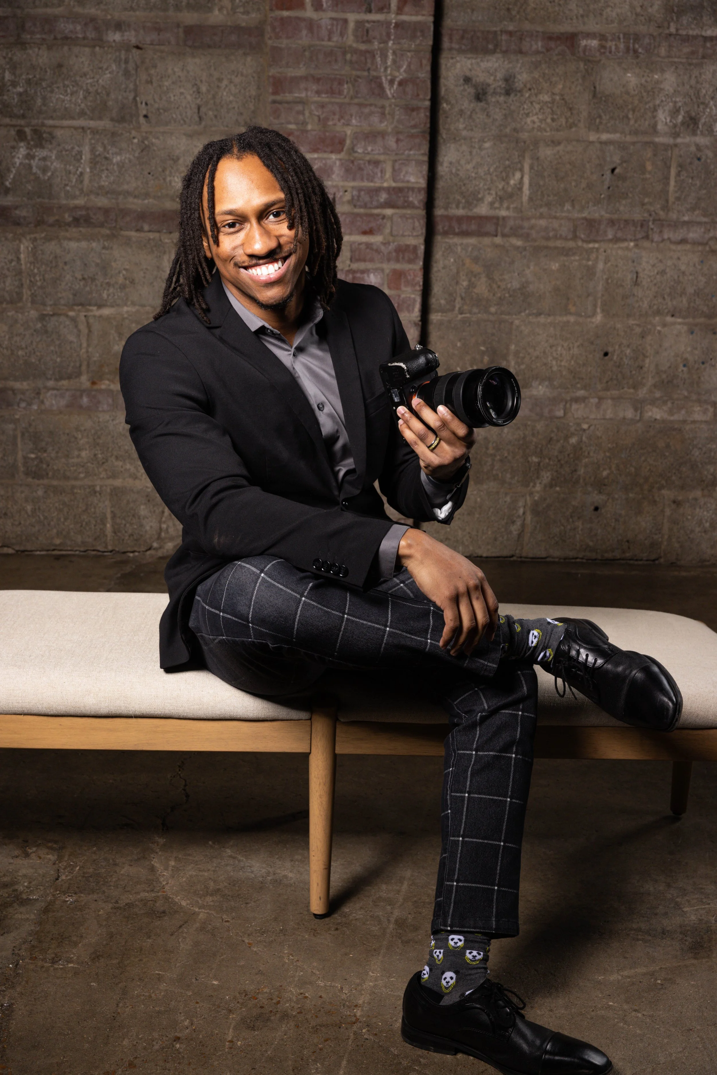 A man with dreadlocks sitting on a beige bench, holding a camera, wearing a black blazer, gray shirt, plaid pants, black shoes, and skull-patterned socks, smiling at the camera against a brick wall background.