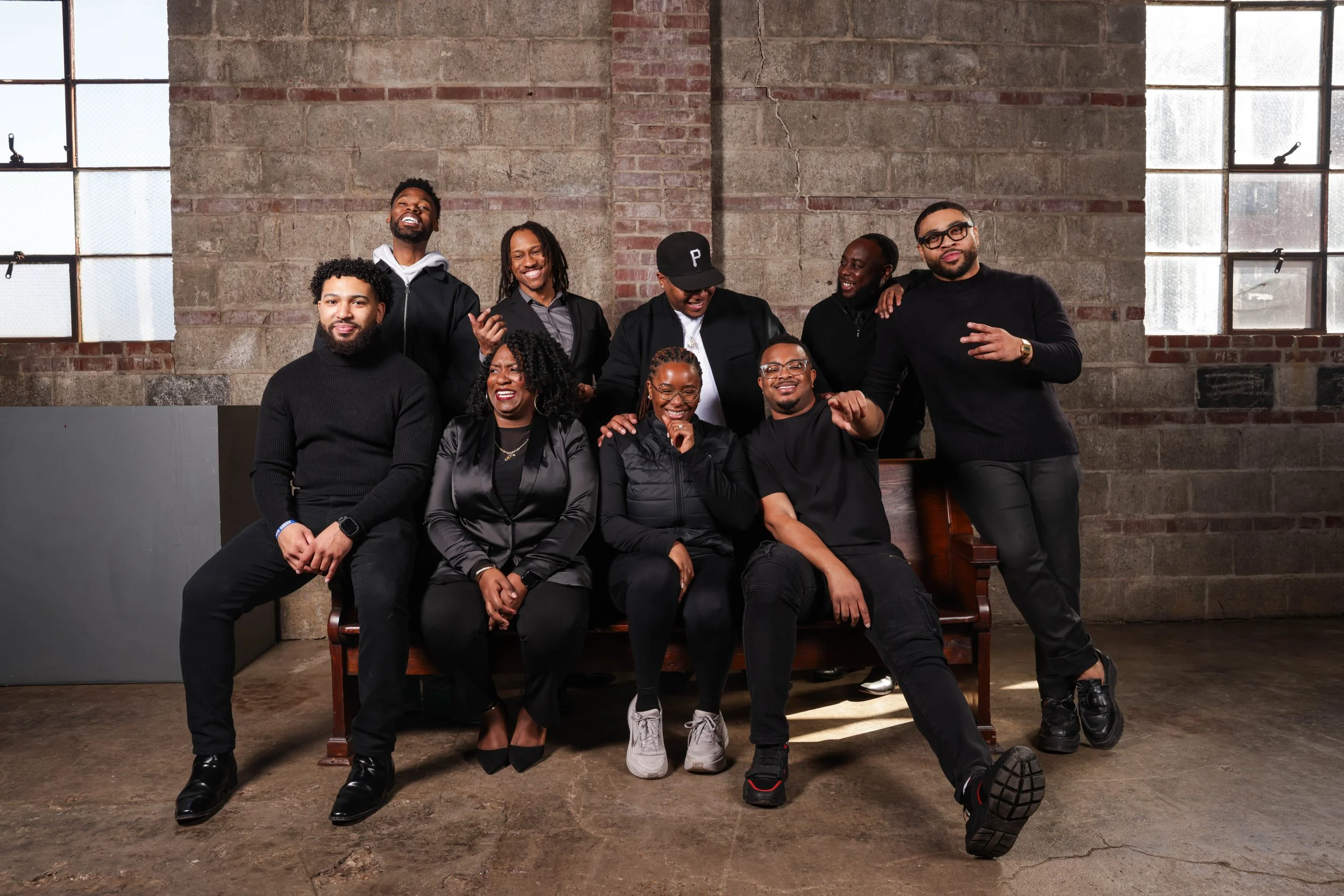 Group of nine diverse people in casual and business attire posing and smiling together in a loft-style space with brick walls and large windows.