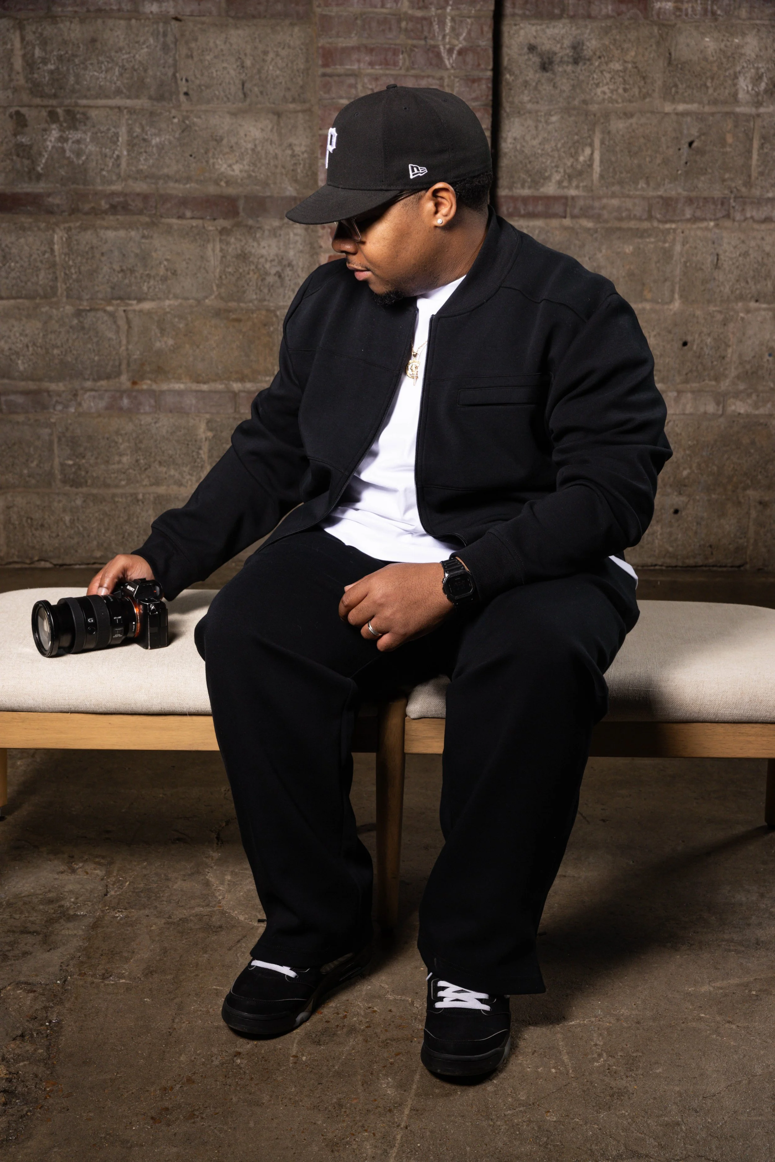 A man sitting on a bench holding a camera, dressed in black streetwear, wearing a black cap, sunglasses, and a watch, with a brick wall background.