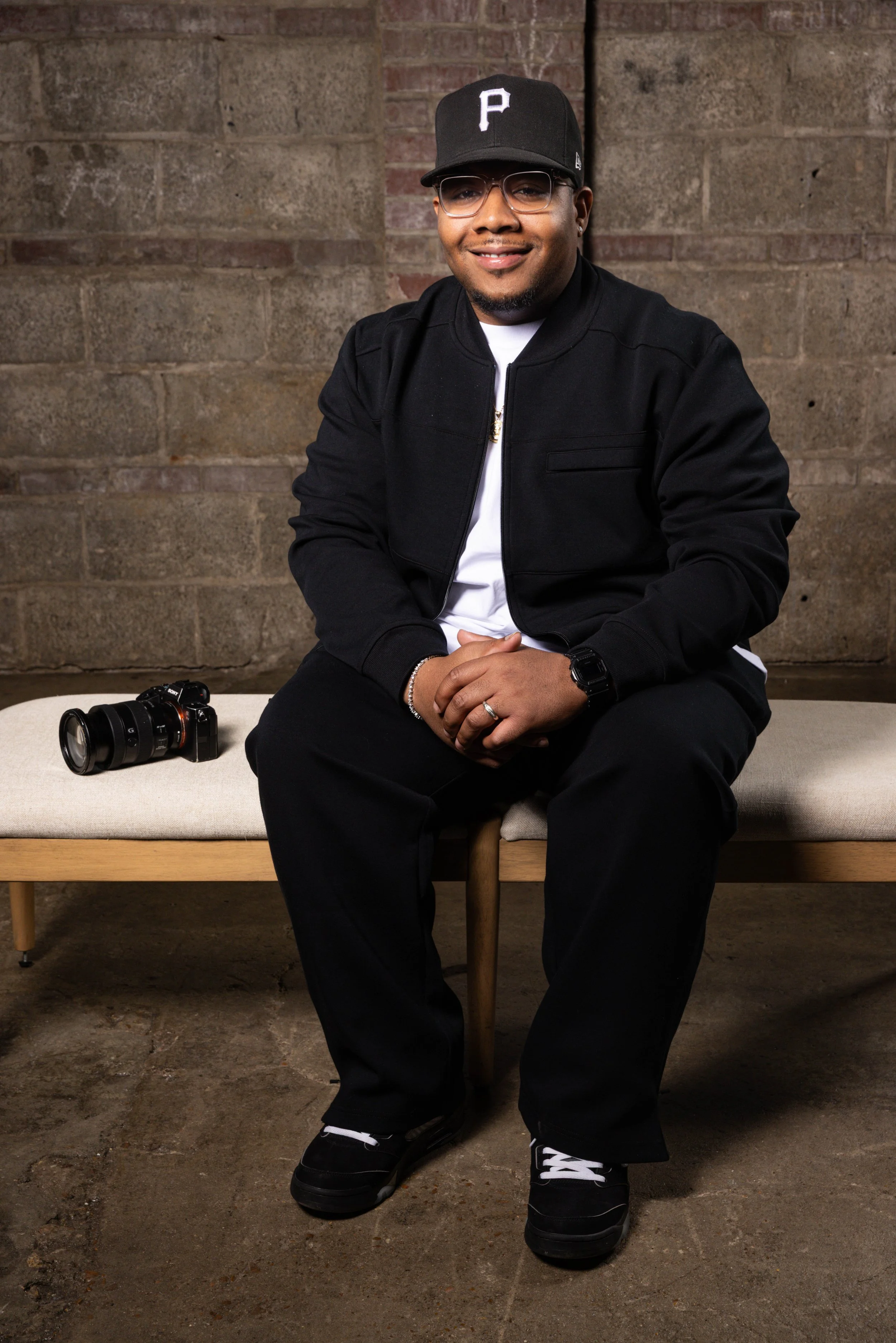 A man sitting on a bench with a camera placed beside him, wearing a black jacket, black pants, black sneakers, a black cap with a white P logo, and glasses, against a brick wall background.