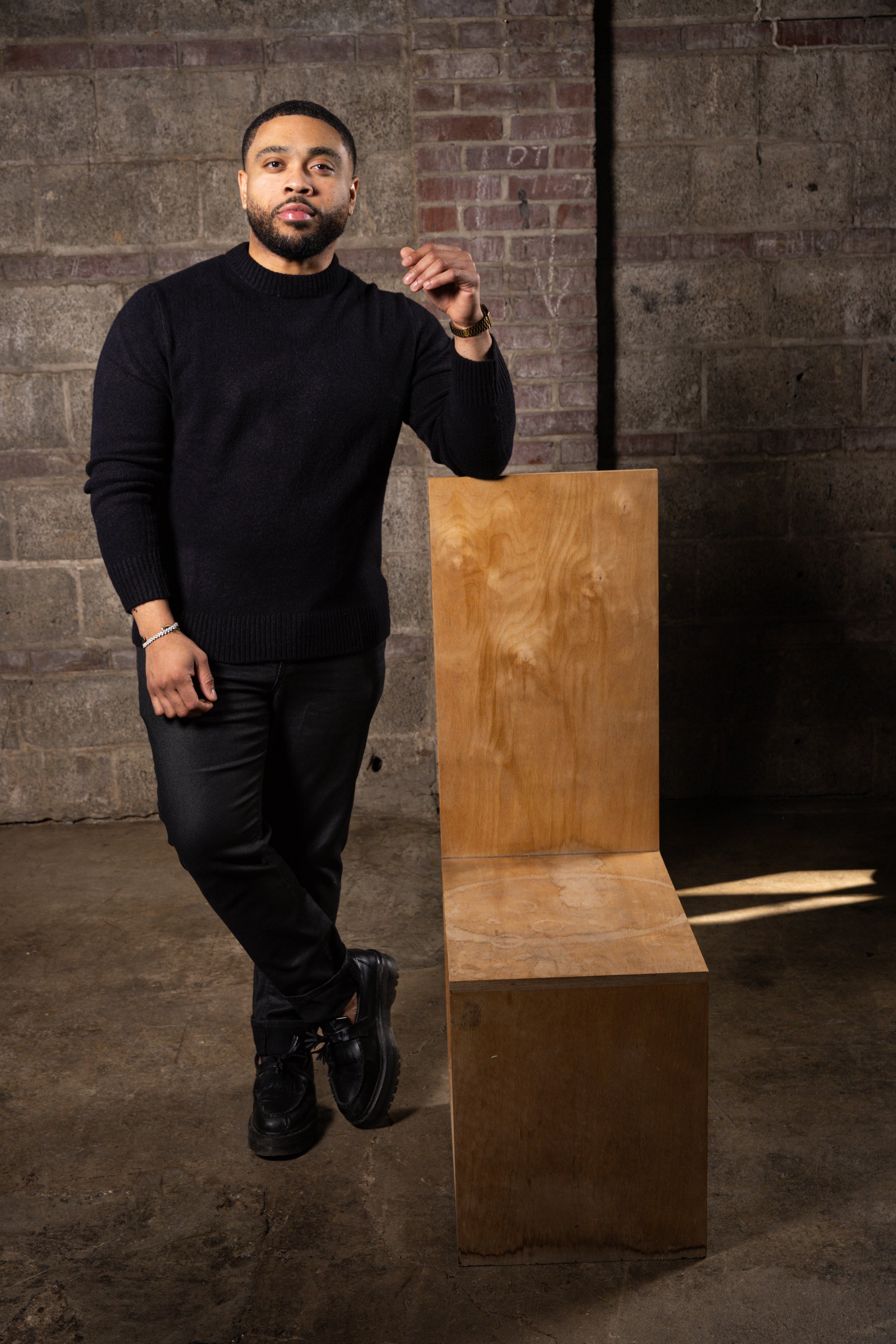 A man with a beard and mustache dressed in a black sweater, black pants, and black shoes leaning against a wooden seat with a brick wall background.