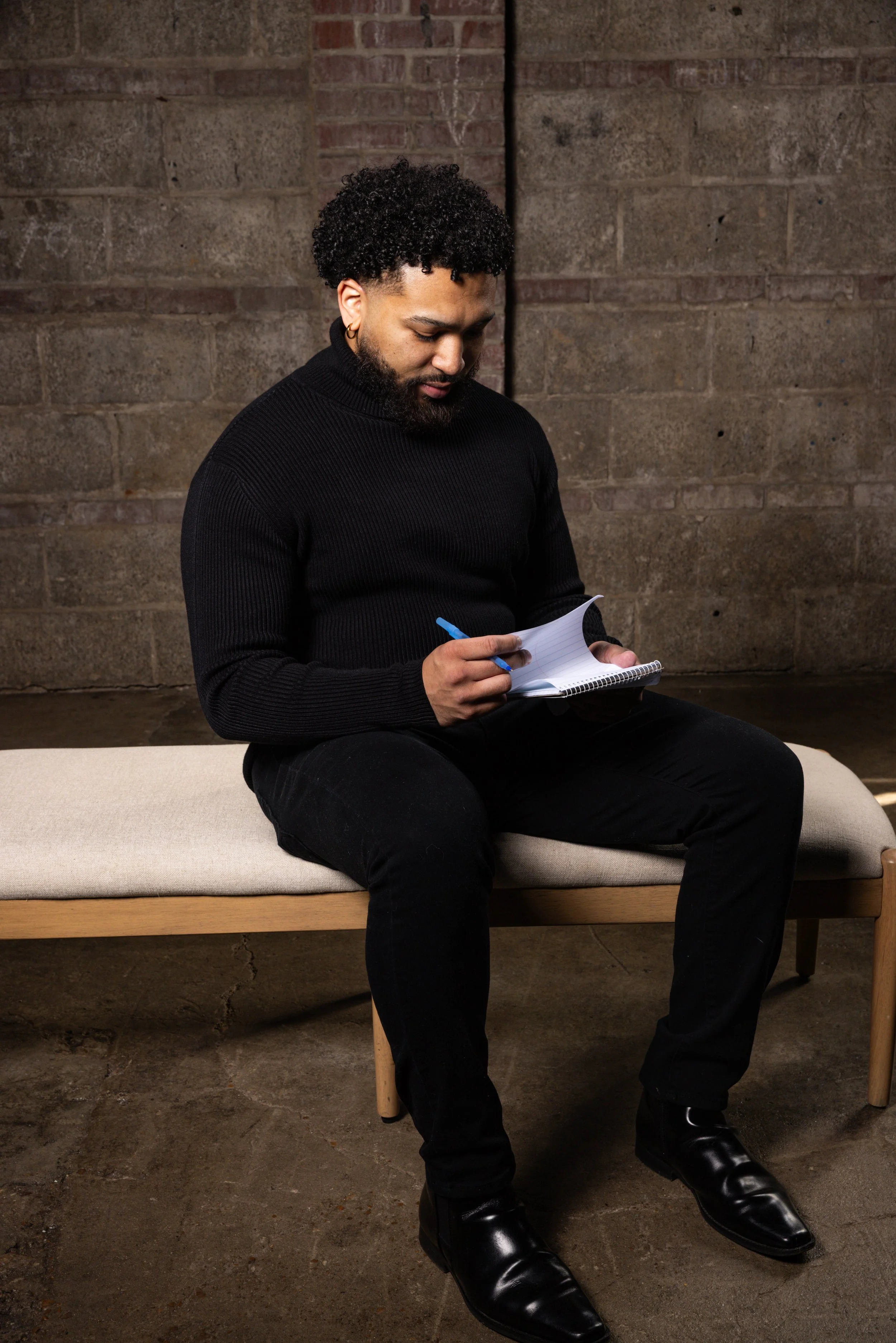 A man with curly hair, beard, and earrings, dressed in black, sitting on a bench with a beige cushion, writing in a notebook against a brick wall background.