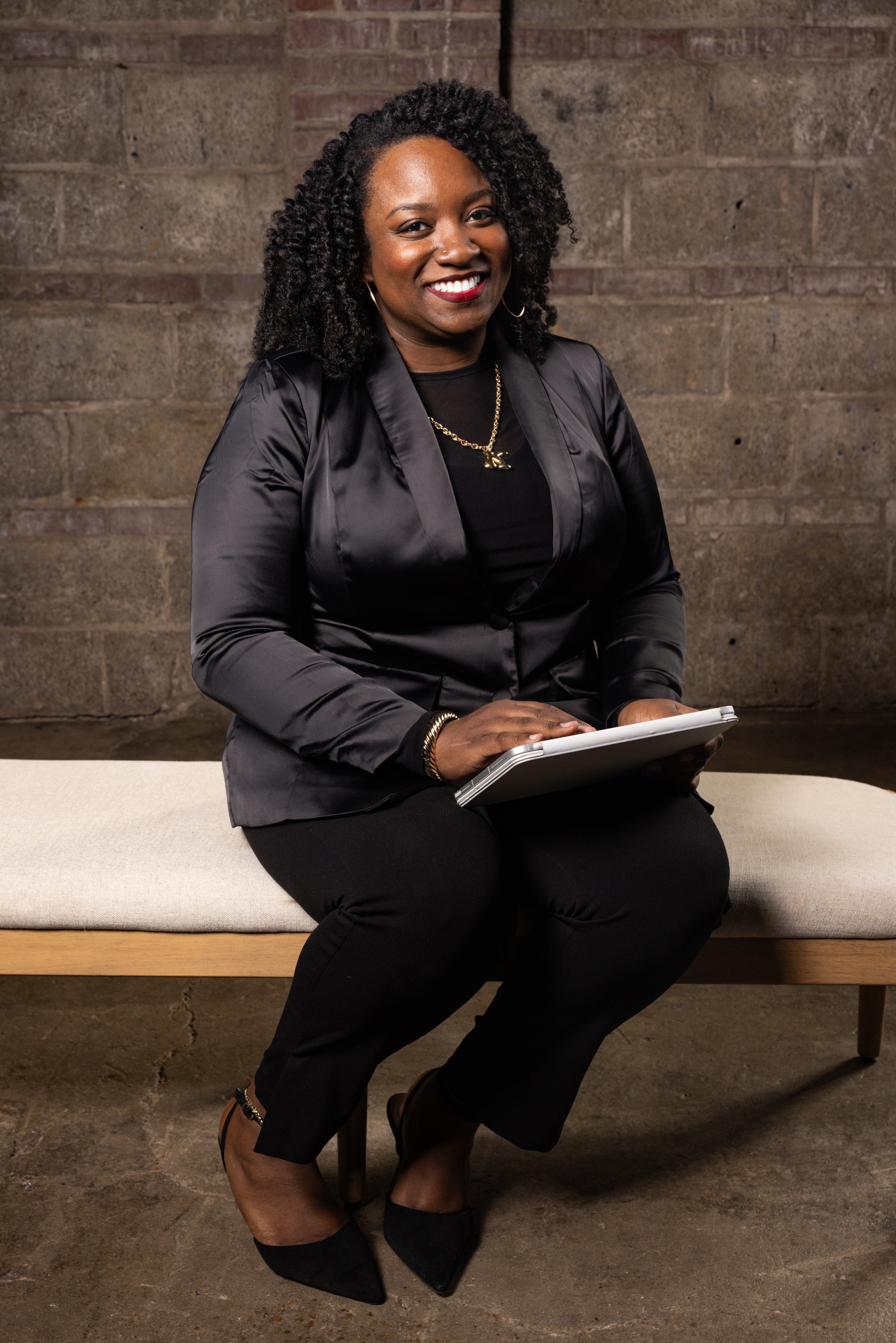 A woman sitting on a bench, smiling, holding a tablet, dressed in a black satin jacket, black pants, and black heels, against a brick wall background.
