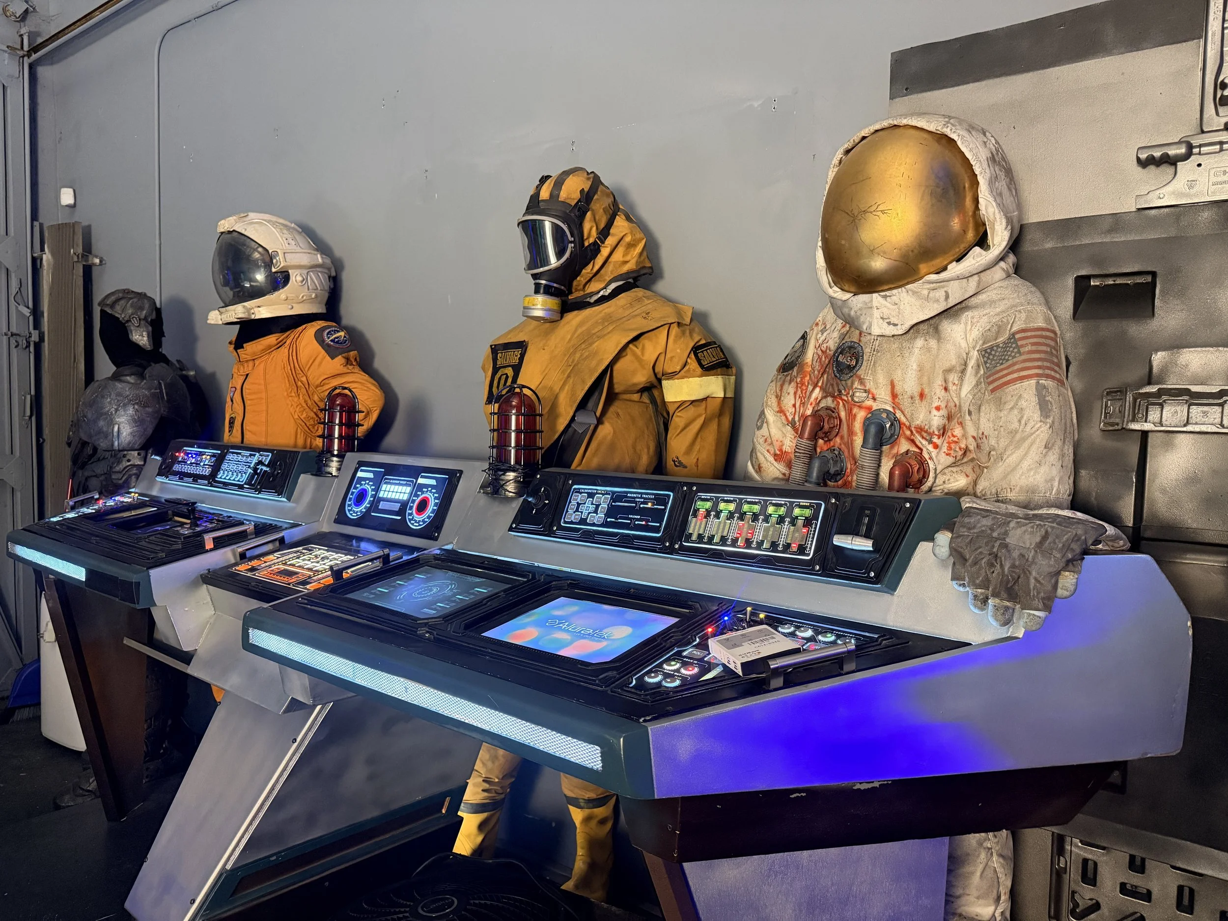 Museum display of four space suits and a control panel from a spacecraft.