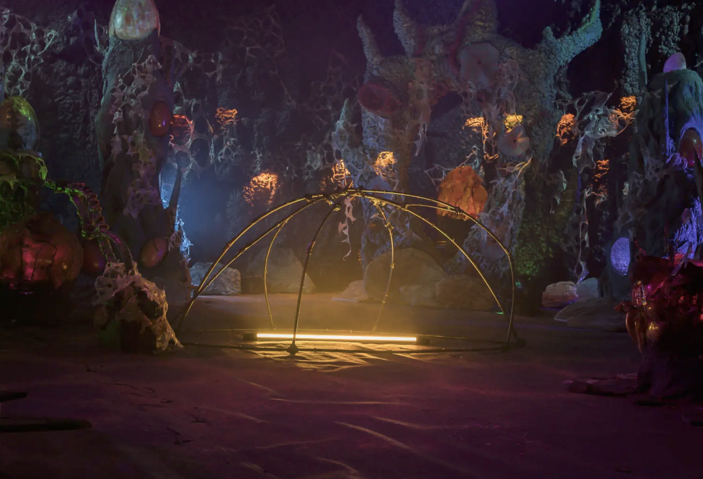 A colorful, illuminated art installation in a dark, cavern-like space featuring organic shapes and glowing sculptures, with a metal dome structure in the center and ambient multicolored lights.