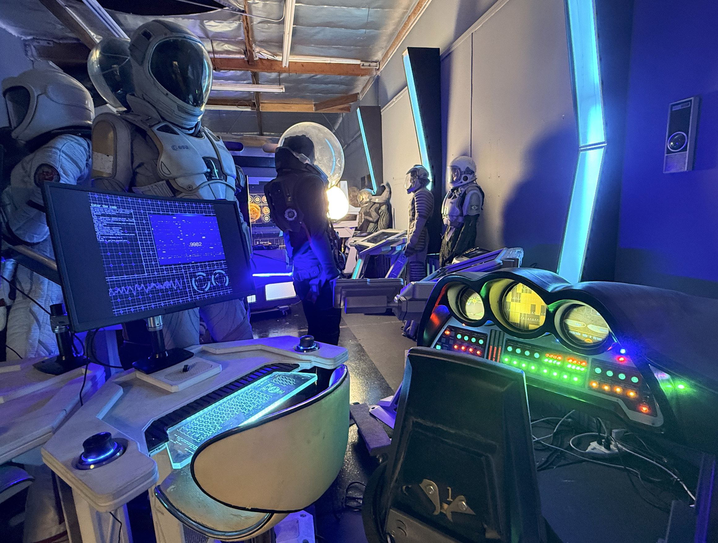 Room with astronaut mannequins, futuristic technology, and colorful neon lights, resembling a sci-fi or space-themed exhibit or arcade.