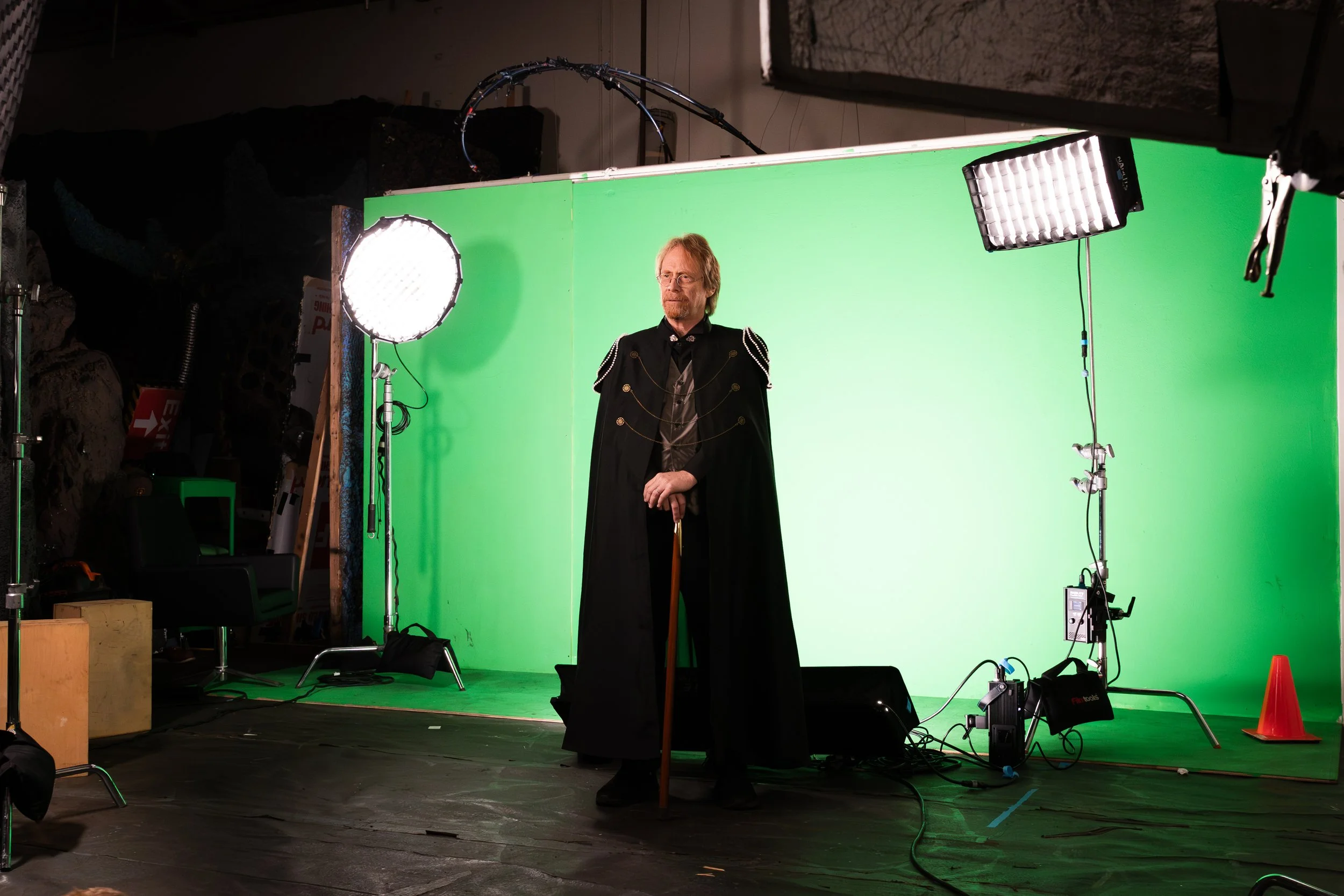 A man dressed in a medieval style black cloak with gold embellishments, holding a cane, standing in front of a green screen in a studio with professional lighting equipment.