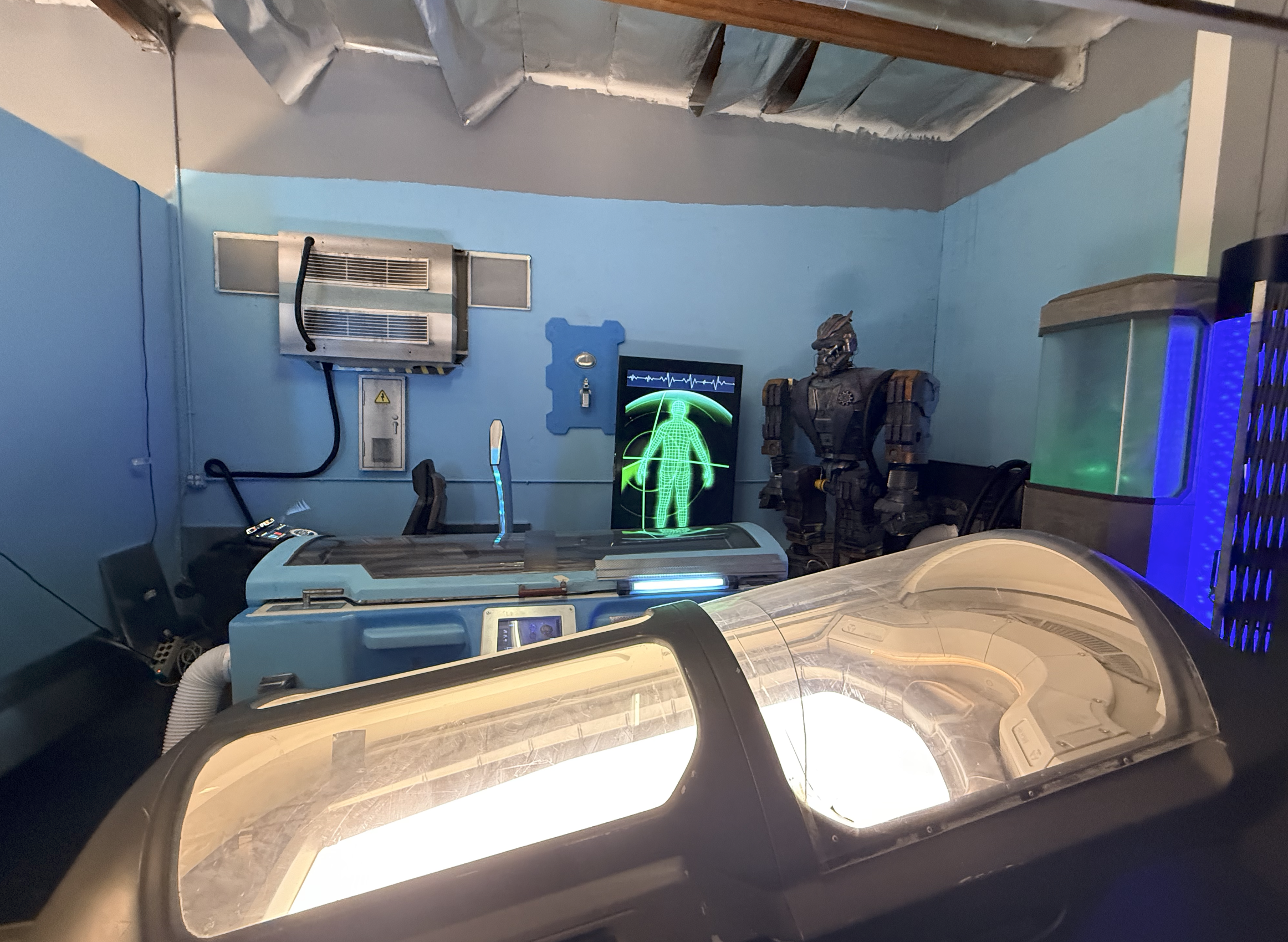 Futuristic medical or examination room with a high-tech incubator in the foreground, a holographic medical scan of a human body on a screen, a humanoid robot, and sci-fi equipment against blue walls.