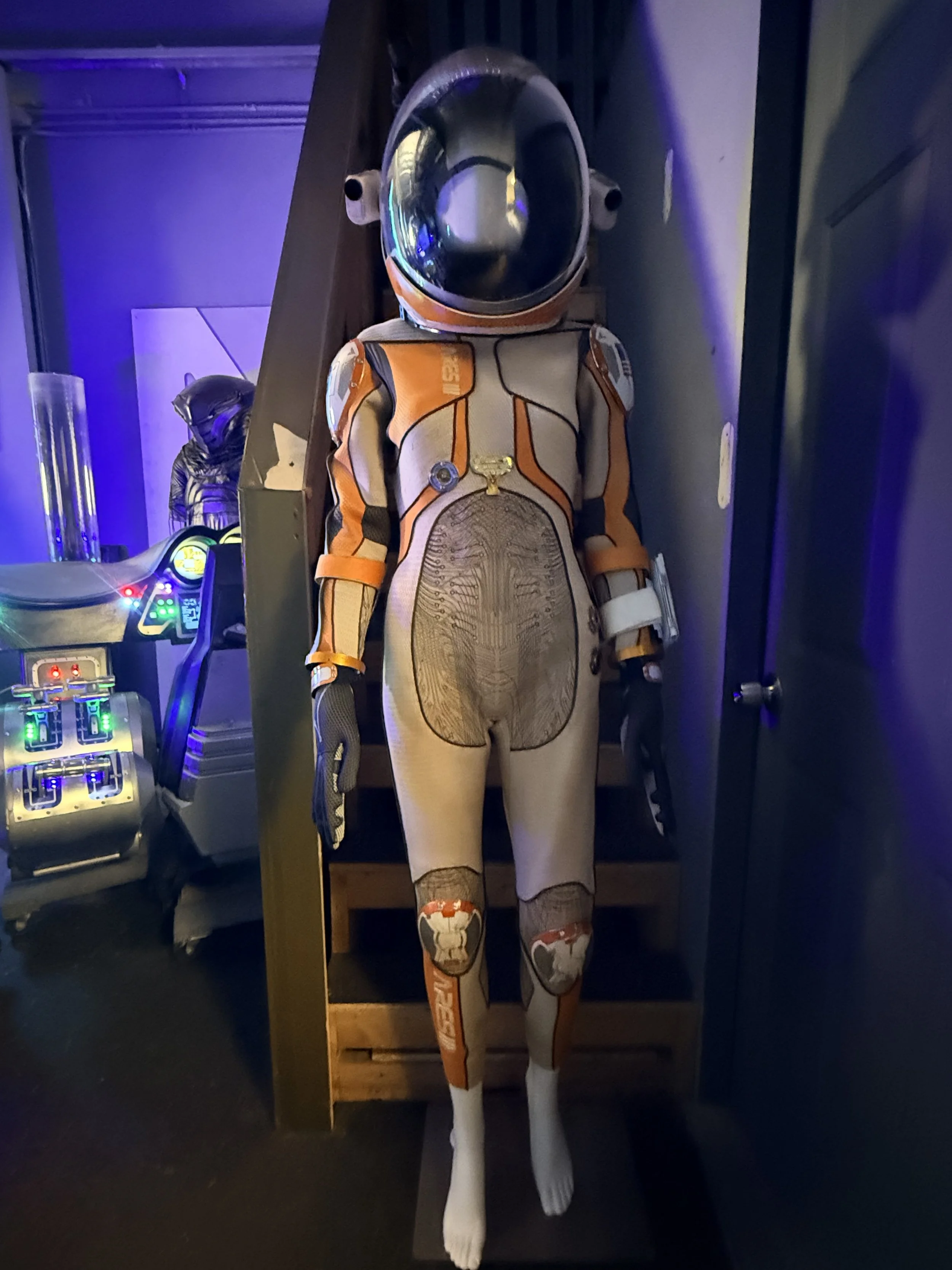 A mannequin dressed as an astronaut in a space suit with a large reflective helmet, standing in a display with scientific and space-themed equipment around.