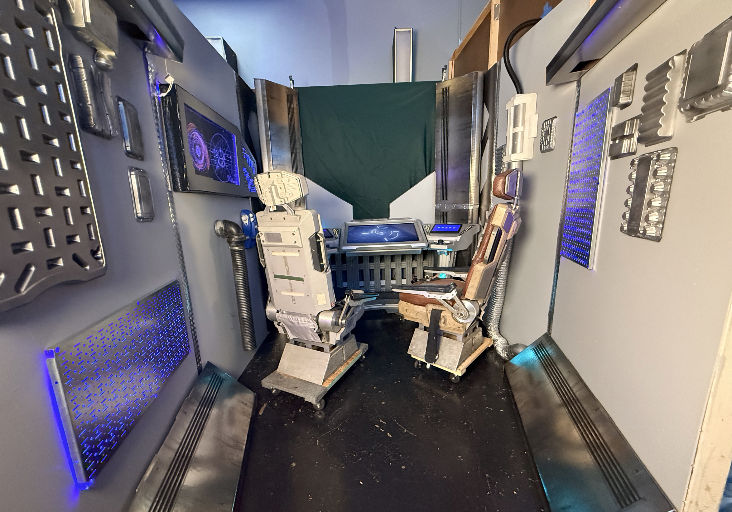 A detailed replica of a spaceship cockpit with two vintage-style seats, control panels, and screens, set against a blue background with a faux window or display area.