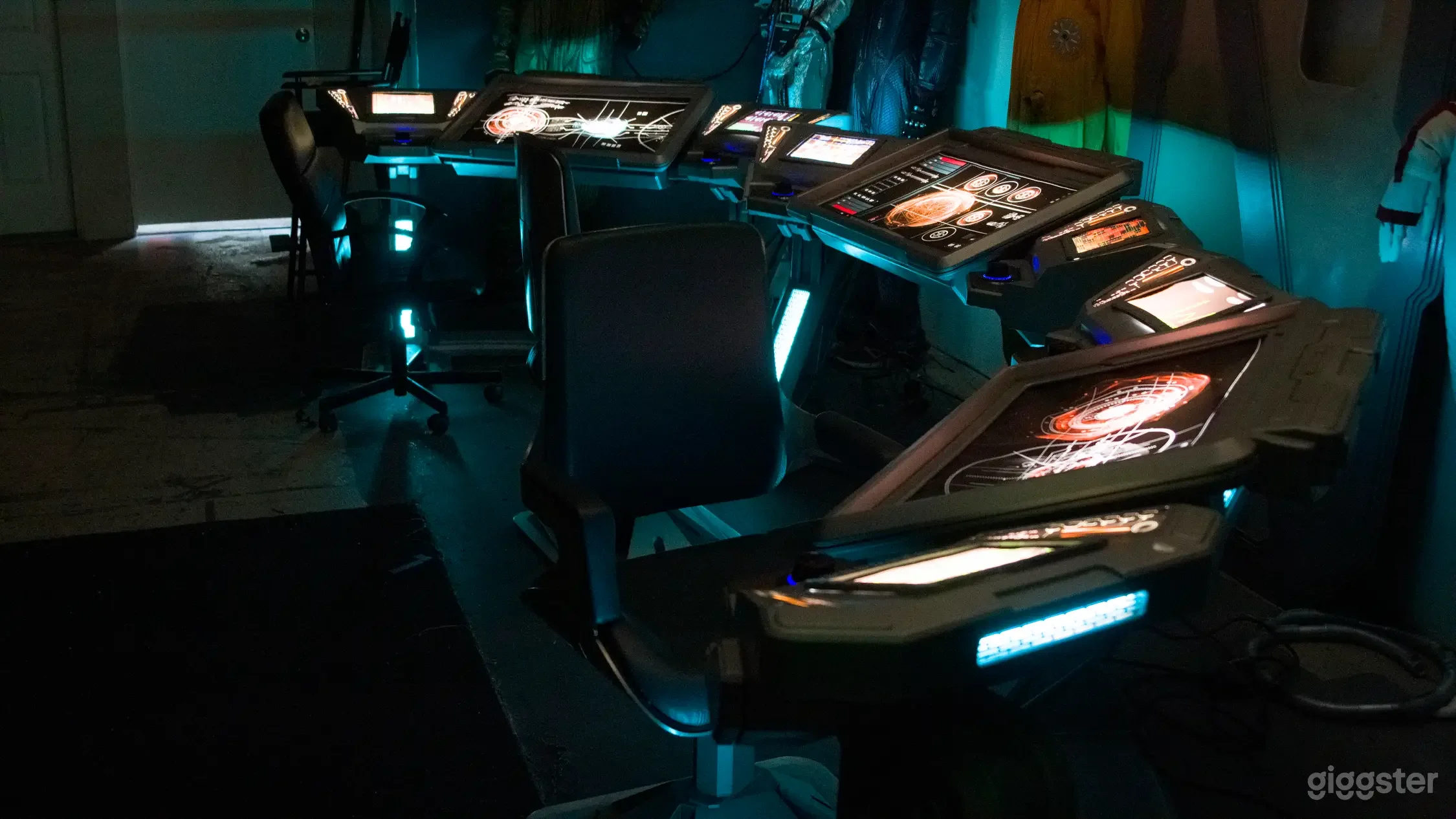 Futuristic control stations with multiple screens displaying intricate graphics, set in a dark room with blue and green lighting.