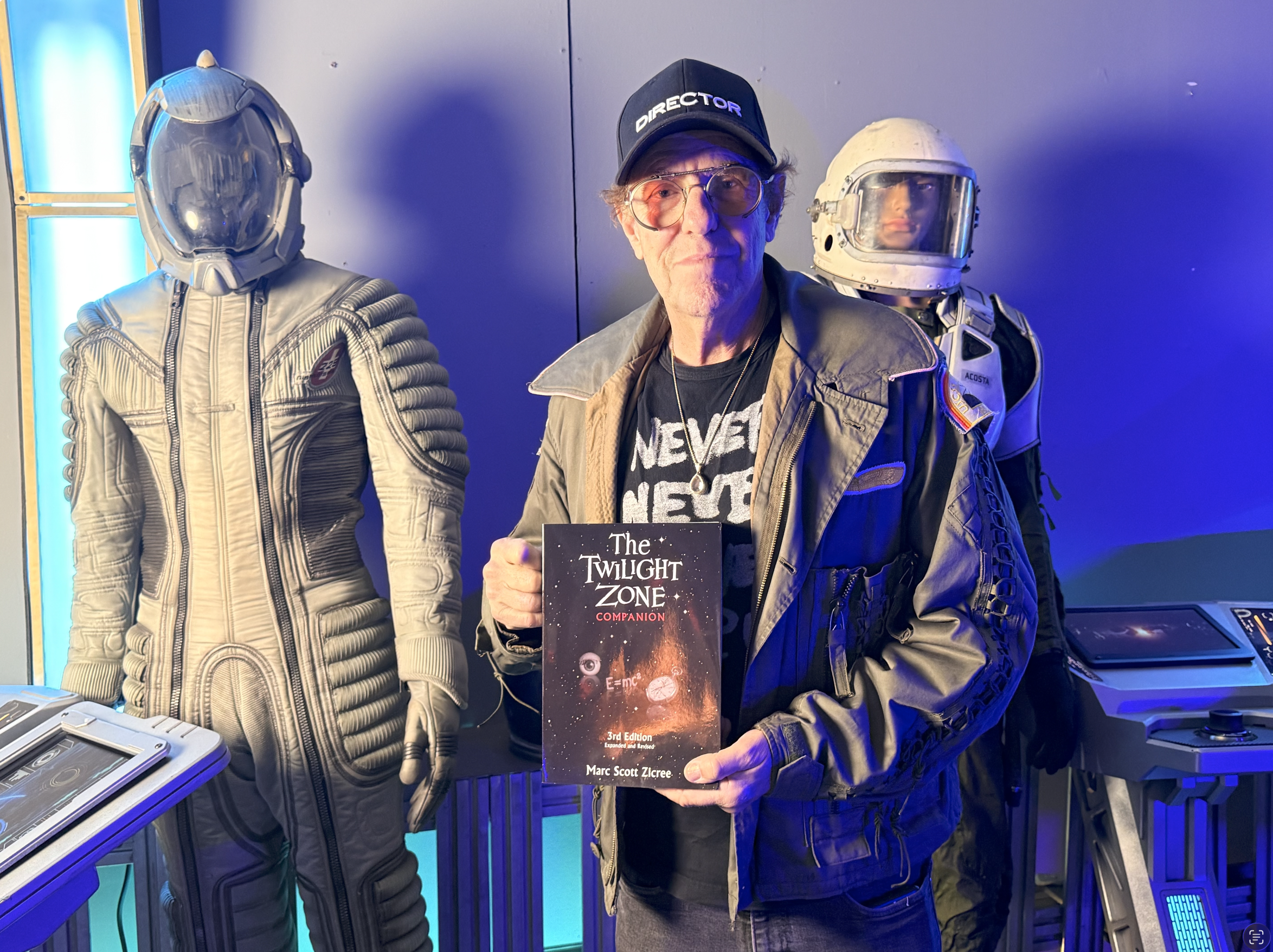 A man holding a book titled 'The Twilight Zone Companion' stands between two astronaut costumes, one with a helmet and the other with a full suit. The man wears glasses, a black cap with 'DIRECTOR' on it, and a brown jacket.