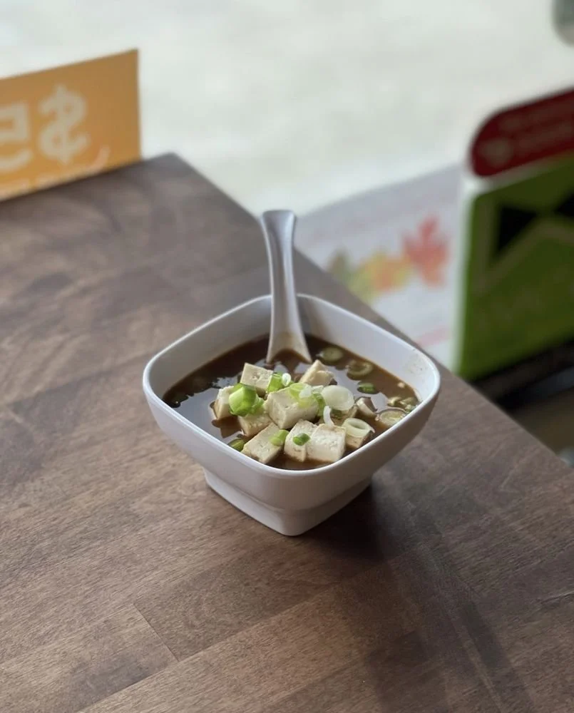A white bowl of hot Chinese-style soup containing tofu, chopped green onions, and soy sauce on a wooden table with a porcelain spoon inside.