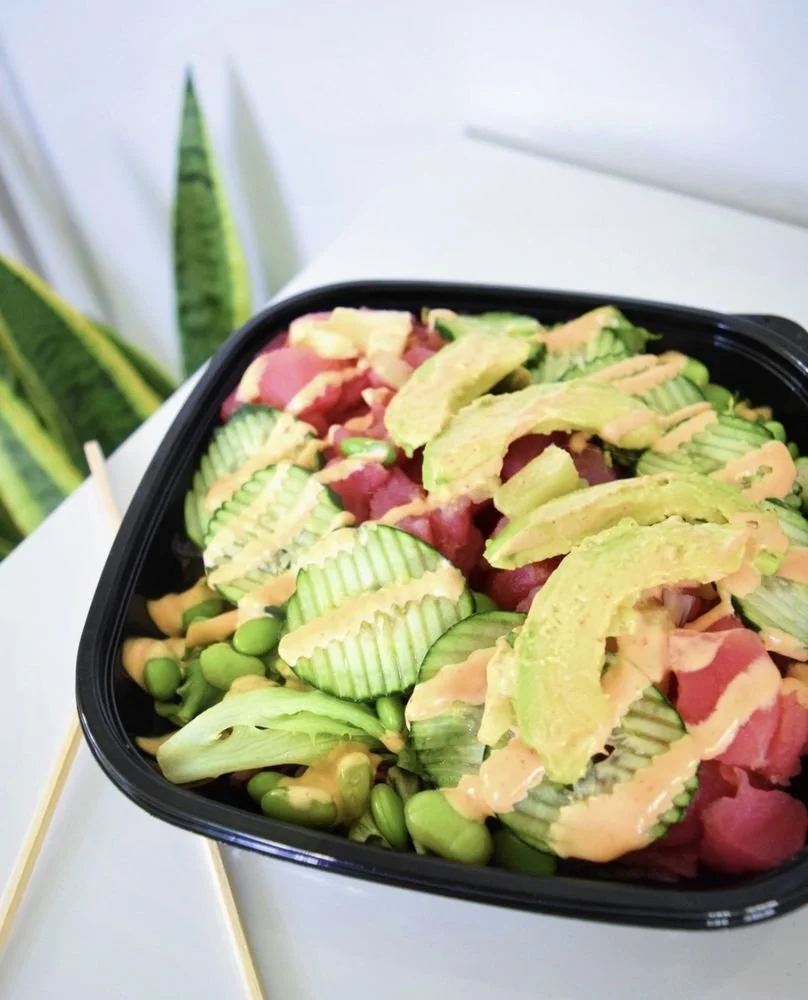 A black container of poke bowl with sliced cucumbers, avocado, salmon, edamame, and drizzled dressing, on a white surface with green plants in the background.