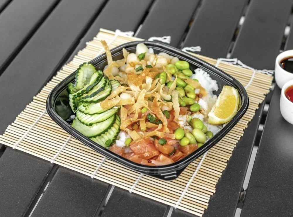 A black takeout container on a bamboo placemat contains a poke bowl with sliced cucumbers, edamame, shredded fried onions, rice, and diced salmon with a lemon wedge on the side. Two small bowls of soy sauce and sriracha are nearby.