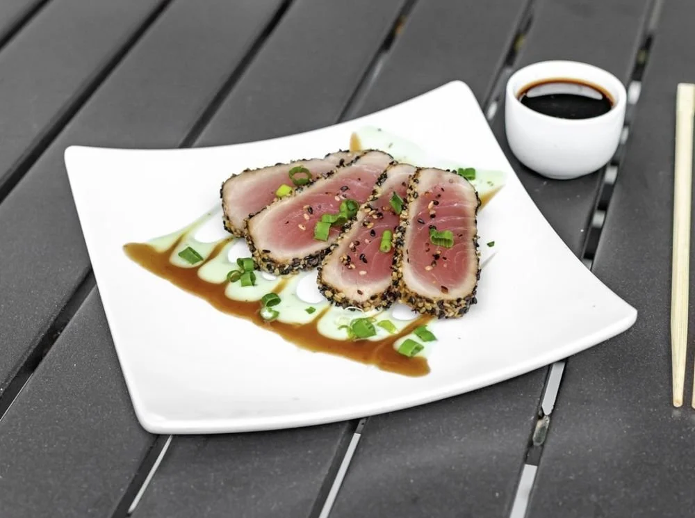 Seared tuna with sesame seeds, green onions, drizzled sauce, and a side of soy sauce on a white plate.