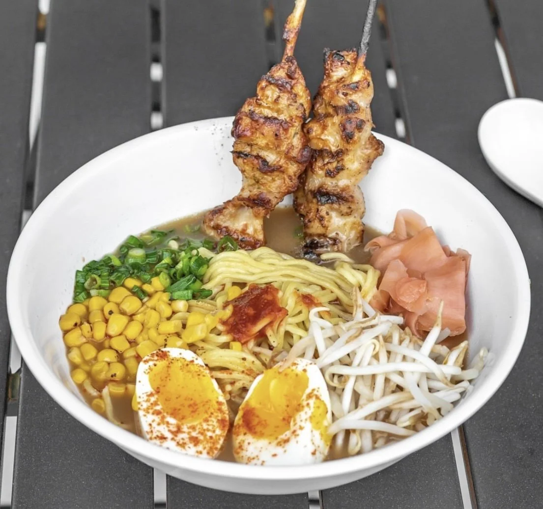Bowl of ramen with grilled chicken skewers, soft-boiled egg halves, corn, green onions, bean sprouts, and slices of pickled ginger on a dark table.