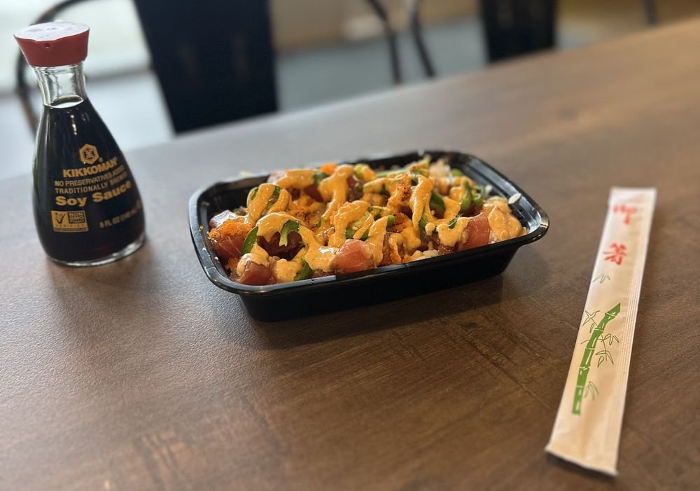 A black plastic container of sushi topped with spicy mayo, a small bottle of soy sauce, and a pair of chopsticks in paper sleeve on a wooden table.