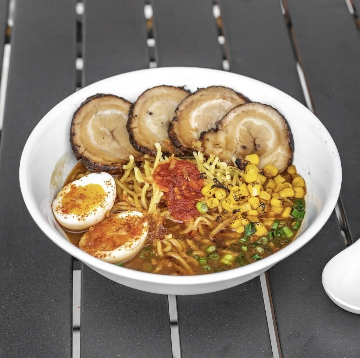 A bowl of ramen noodles topped with slices of pork, a soft boiled egg, corn, chopped green onions, and a dollop of red chili paste.