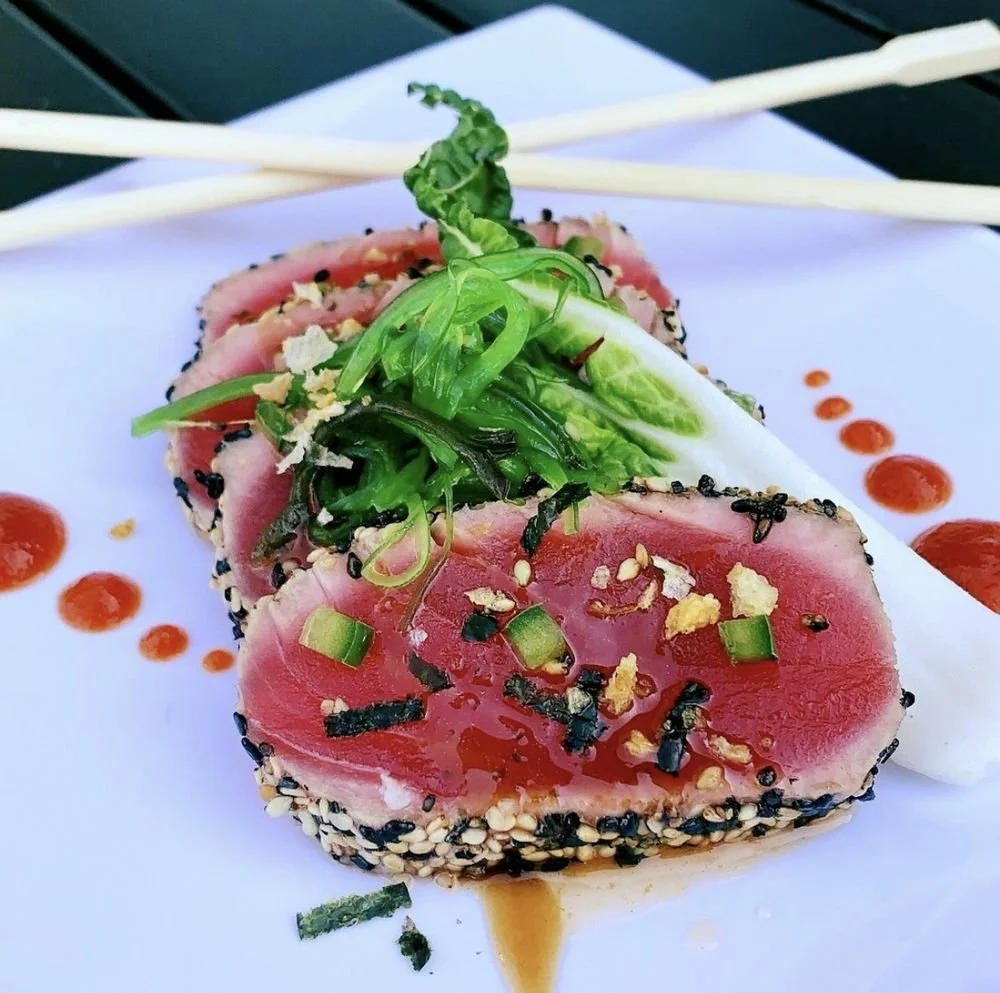 Seared tuna steak coated with sesame seeds topped with green onions and seaweed garnishes on a white plate, with soy sauce and hot sauce drizzle, accompanied by white chopsticks.