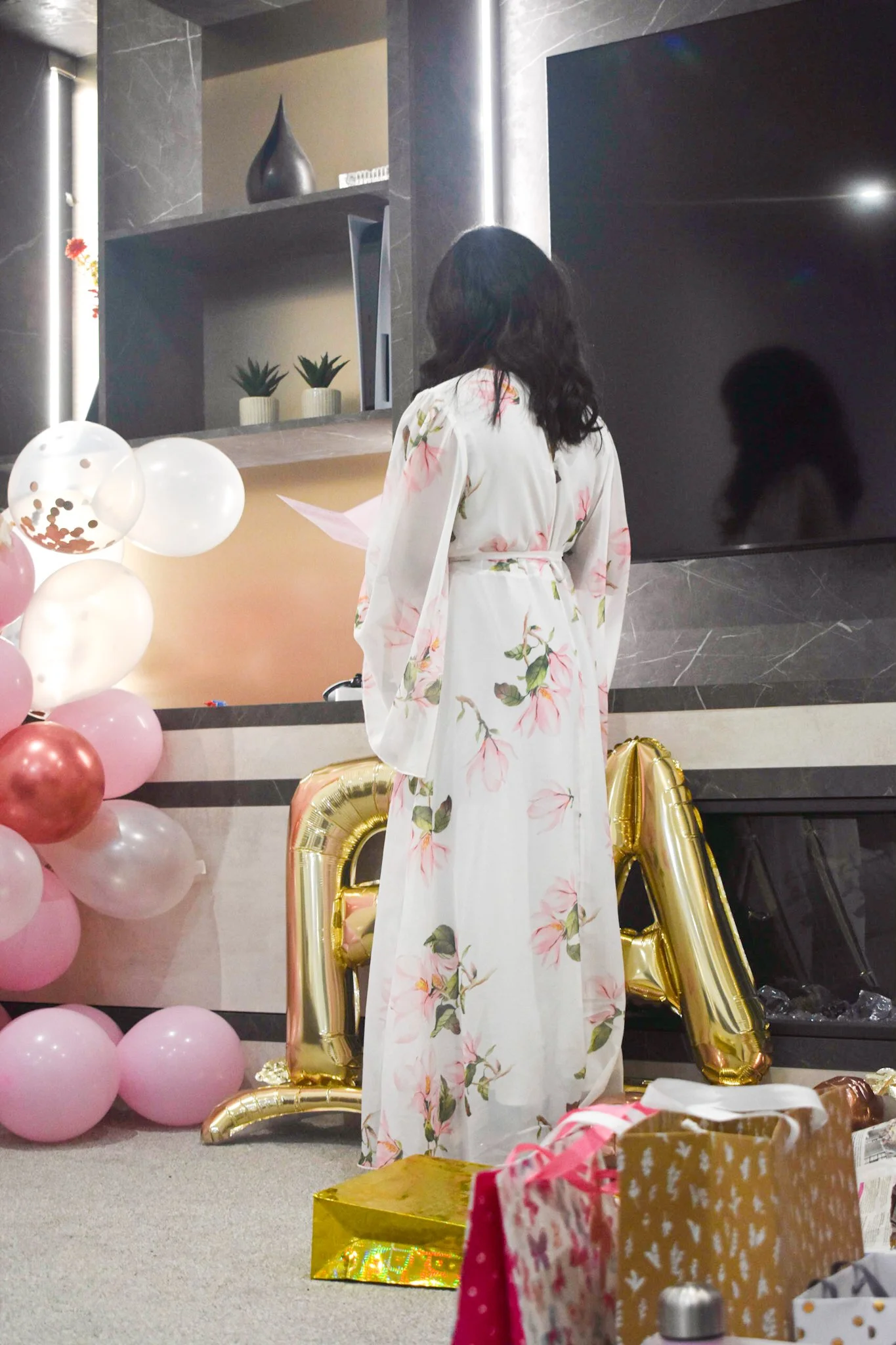A woman in a long, white floral dress is standing indoors near pink balloons and large gold metallic balloons that spell 'BABY'. She appears to be reading or speaking from a piece of paper, with her back facing the camera. The setting suggests a cele