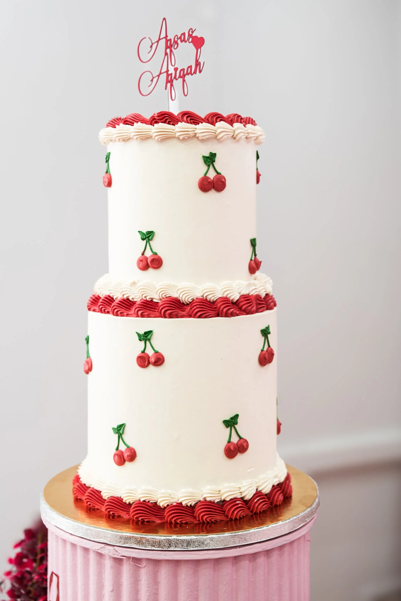A three-tiered wedding cake with white icing, decorated with small cherry and leaf designs, topped with a pink cake topper that says "Azaar Azzaah."
