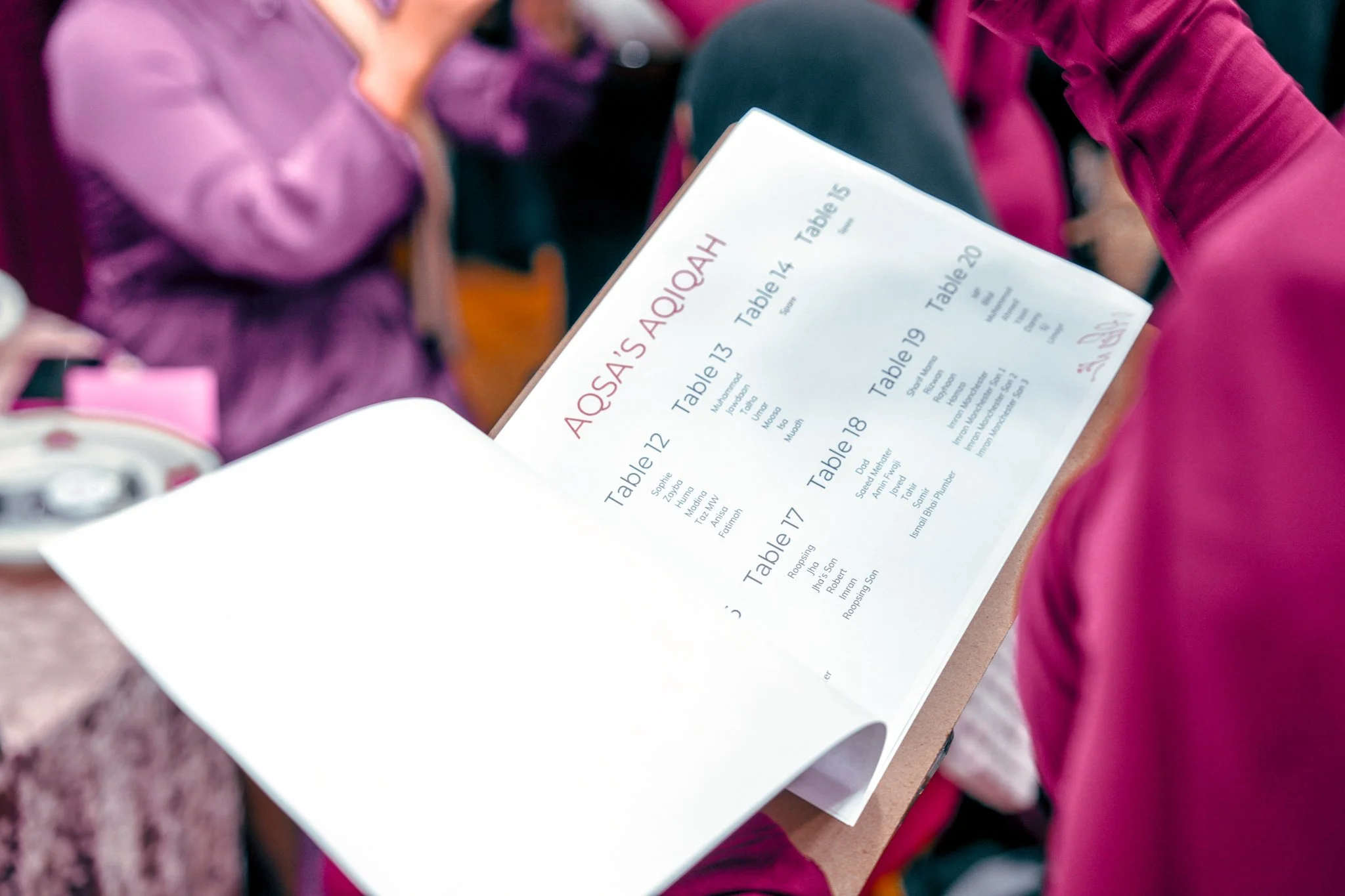 A person holding a printed seating chart for an event, listing tables and names, titled "Aosá’s A😊l 😛ad".