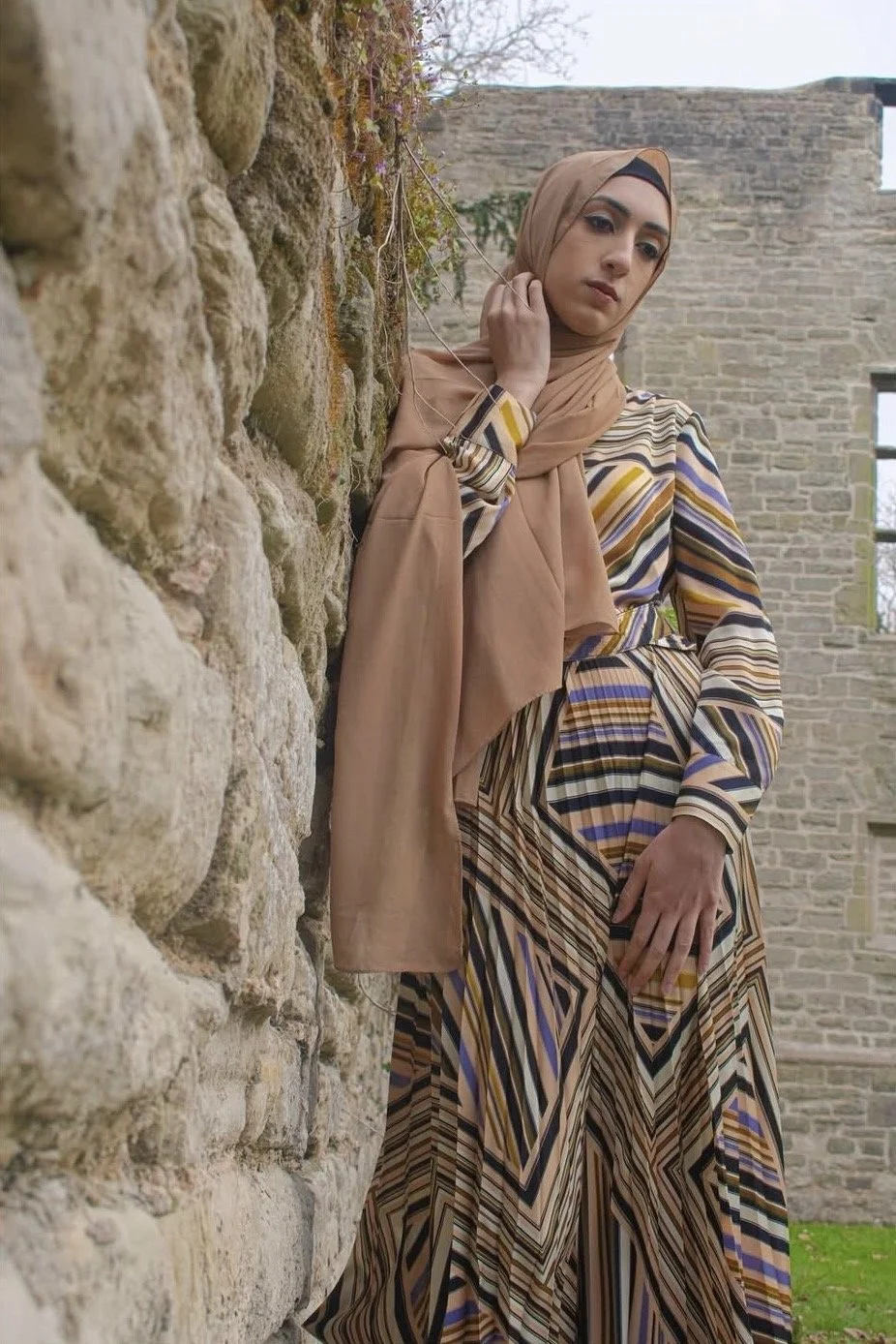 A woman wearing a beige hijab and a long, patterned dress standing against an old stone wall, with green grass and a brick building in the background.