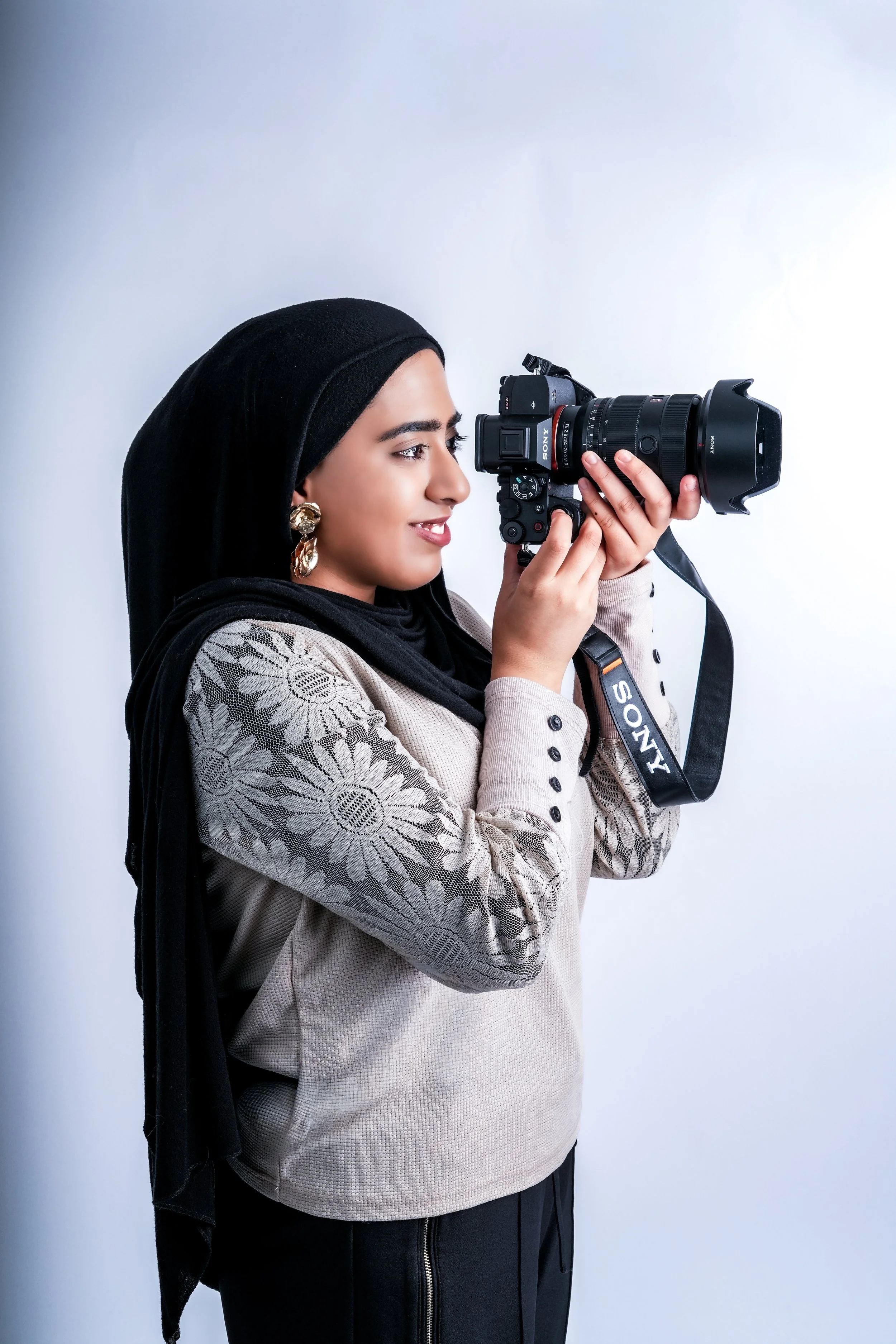 A woman with light brown skin, wearing a black hijab, gold earrings, and a beige top with floral embroidery, is holding and looking through a Sony DSLR camera against a plain white background.