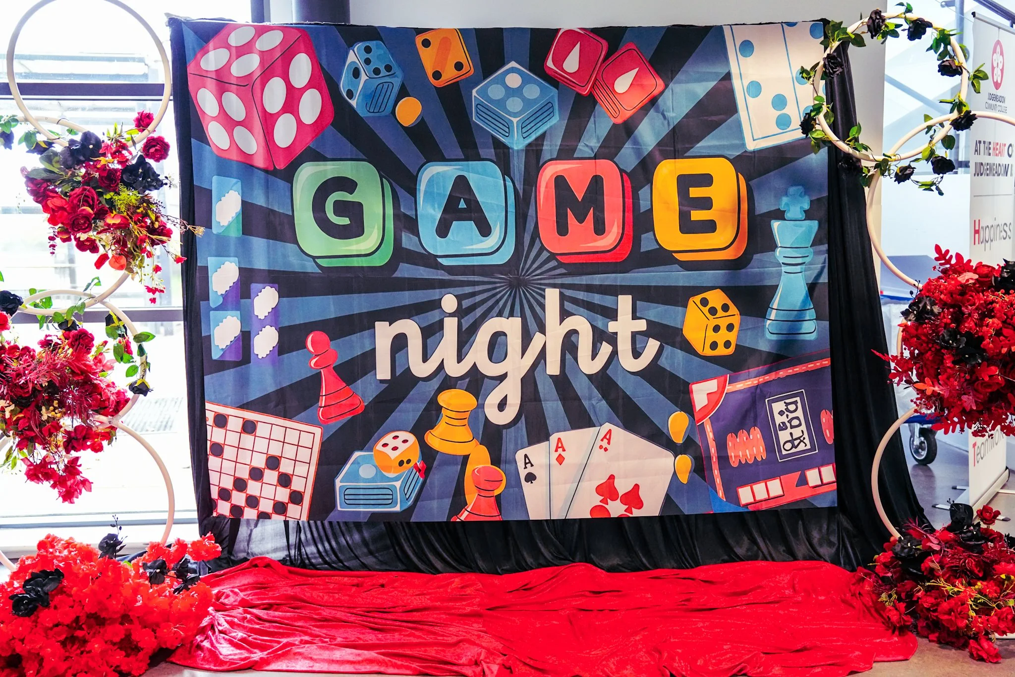 Decorative banner for a game night event, featuring colorful illustrations of dice, chess pieces, playing cards, and board game components, surrounded by floral arrangements and set against a curtain with red fabric on the floor.