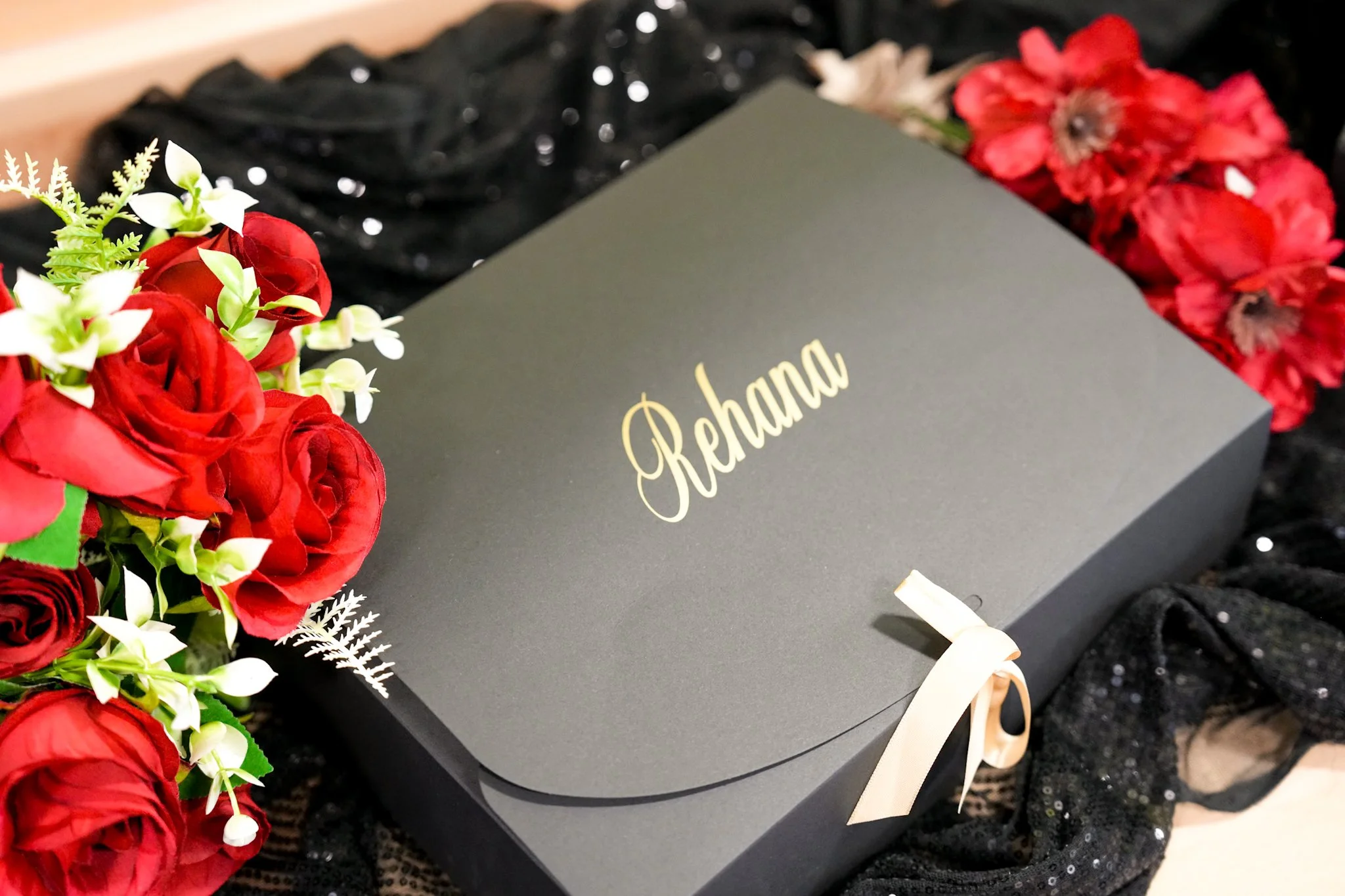 A black gift box with gold text that says 'Rehana' surrounded by red roses, white flowers, and black glittery fabric.