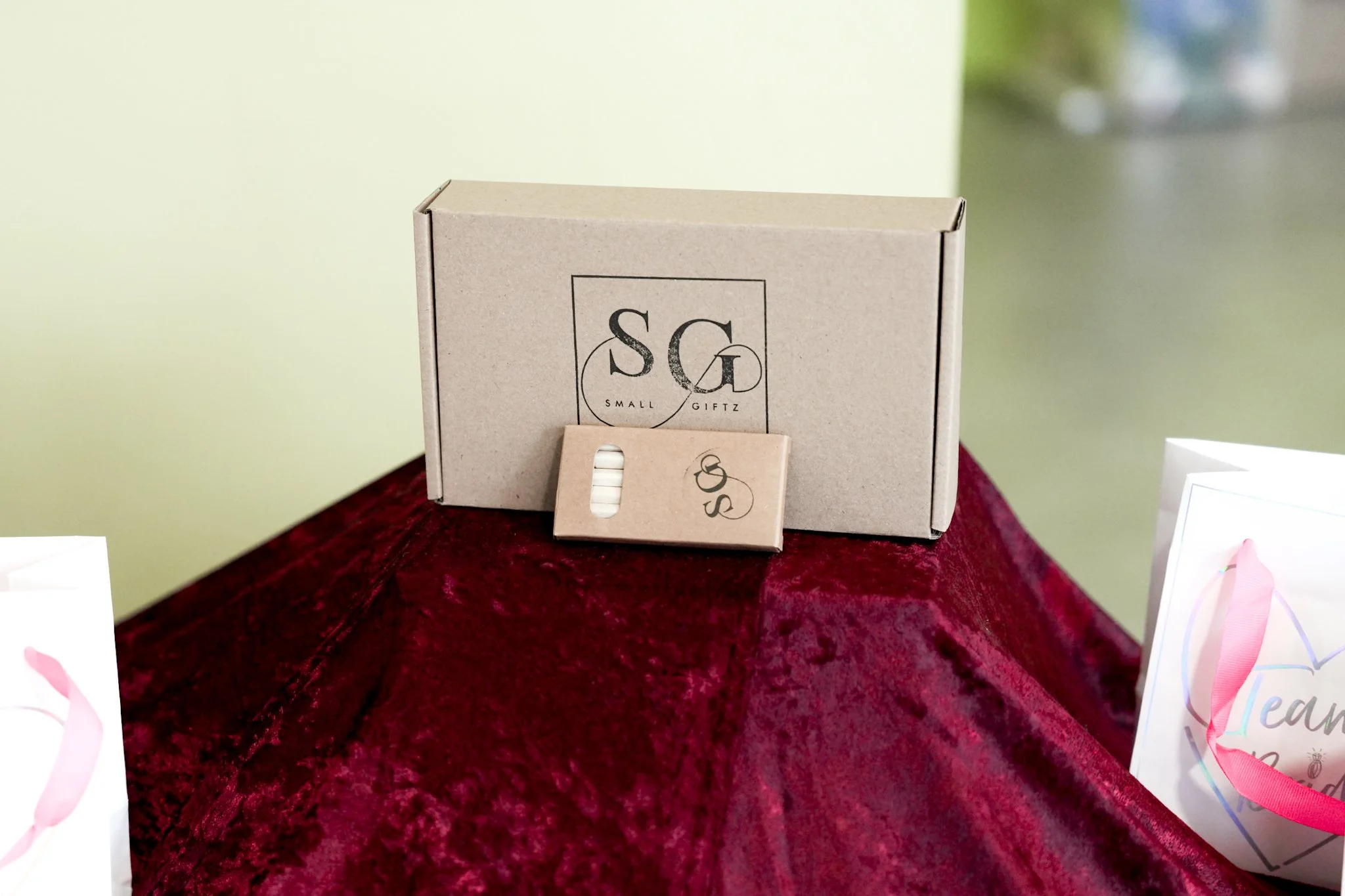 A small beige box labeled "SG Small Giftz" with a smaller matching packet placed in front, sitting on a red velvet surface outdoors with a blurred background.