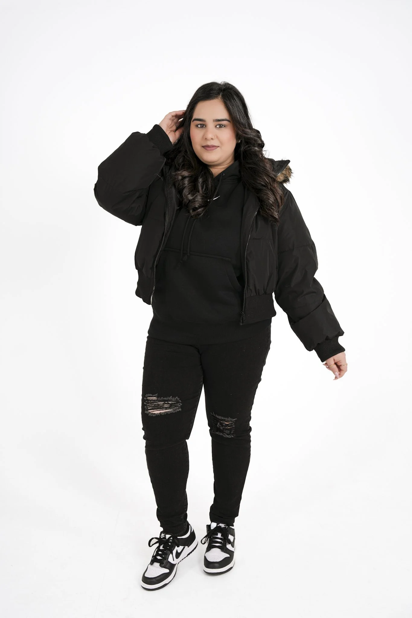 A woman with long dark hair wearing a black quilted jacket, black hoodie, ripped black jeans, and black-and-white Nike sneakers posing against a plain white background.