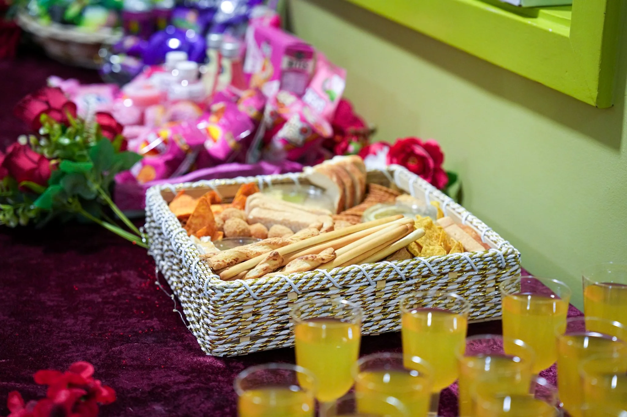 A table with a woven basket of various foods, surrounded by small yellow drinks, flowers, and packaged snacks in the background.