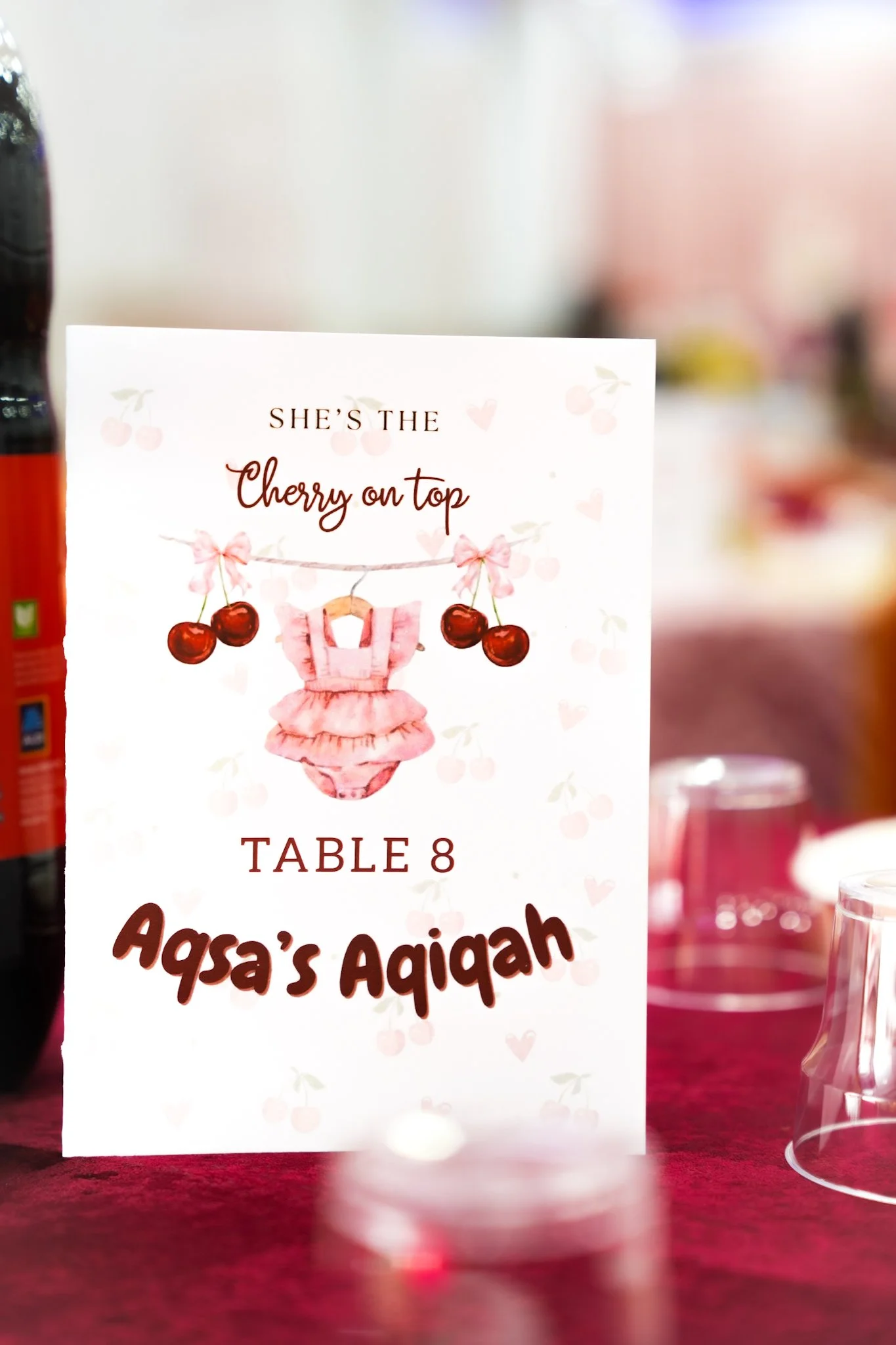Table sign with cherry theme for a girl named Aqsa Aqiqah, displaying her table number 8, featuring a clothesline with cherries and a pink dress.