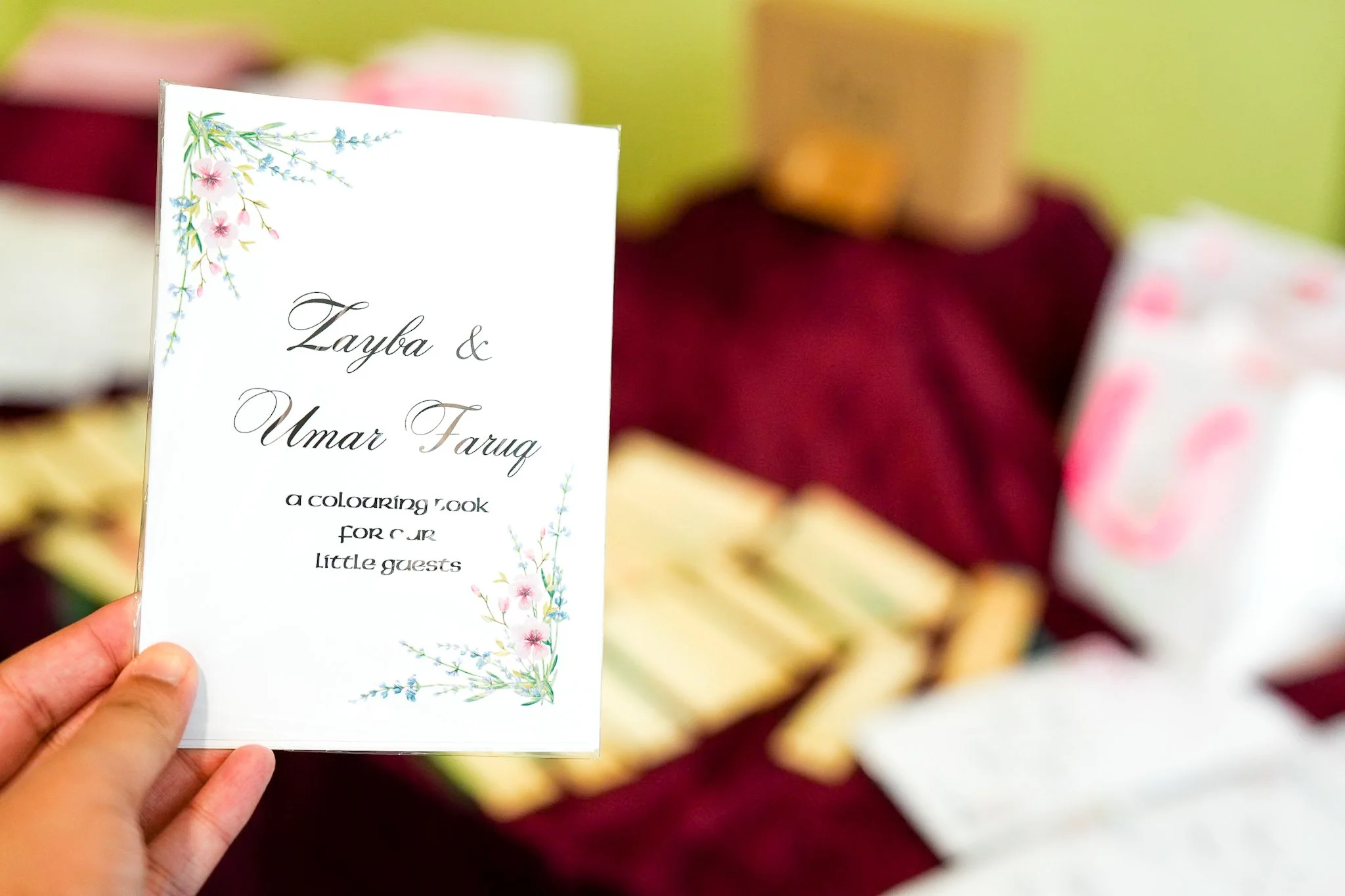 Person holding a white booklet titled 'Zayba & Umar Faruq, a coloring book for our little guests' with floral decorations, with blurred colorful gifts or items in the background.