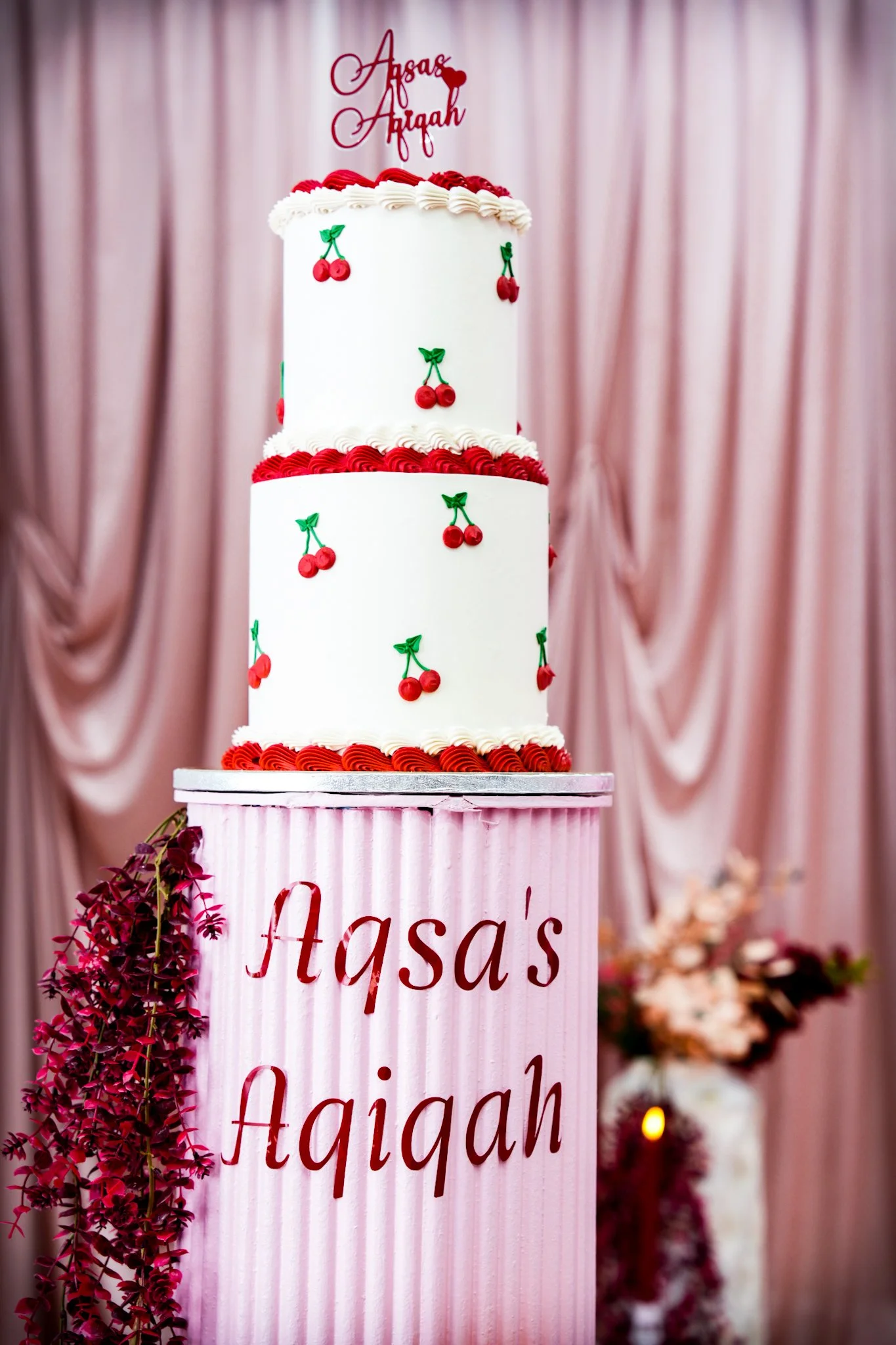 A two-tiered white cake decorated with cherry motifs and red piping, placed on a pink stand with the inscription 'Aqsa Aqiqah,' in front of pink curtains.