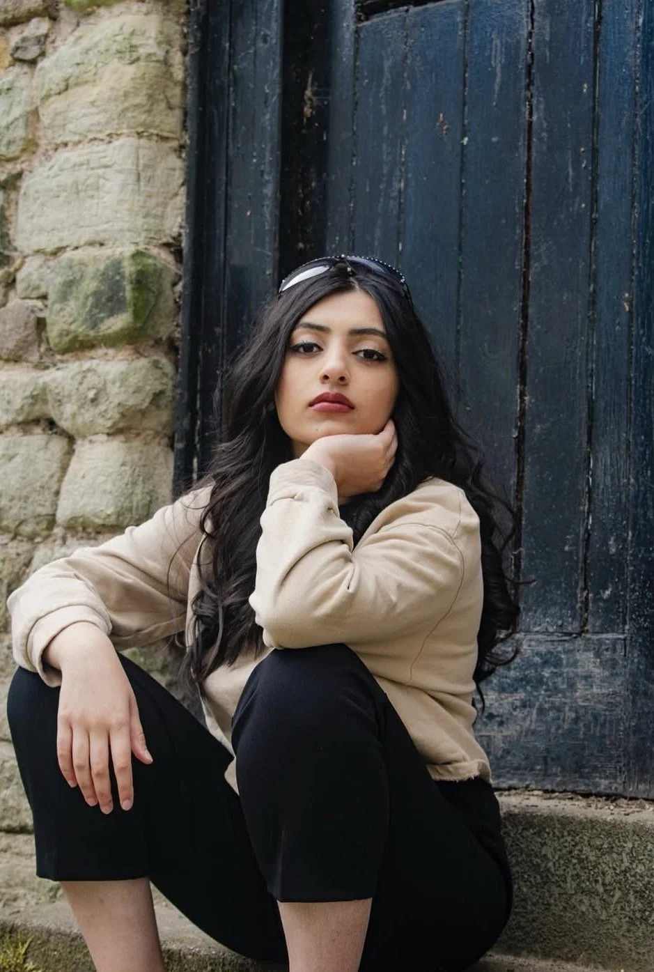 A young woman with long dark hair, wearing a beige jacket and black pants, sitting on the ground with her knees up and one hand supporting her chin, in front of a dark wooden door and stone wall.