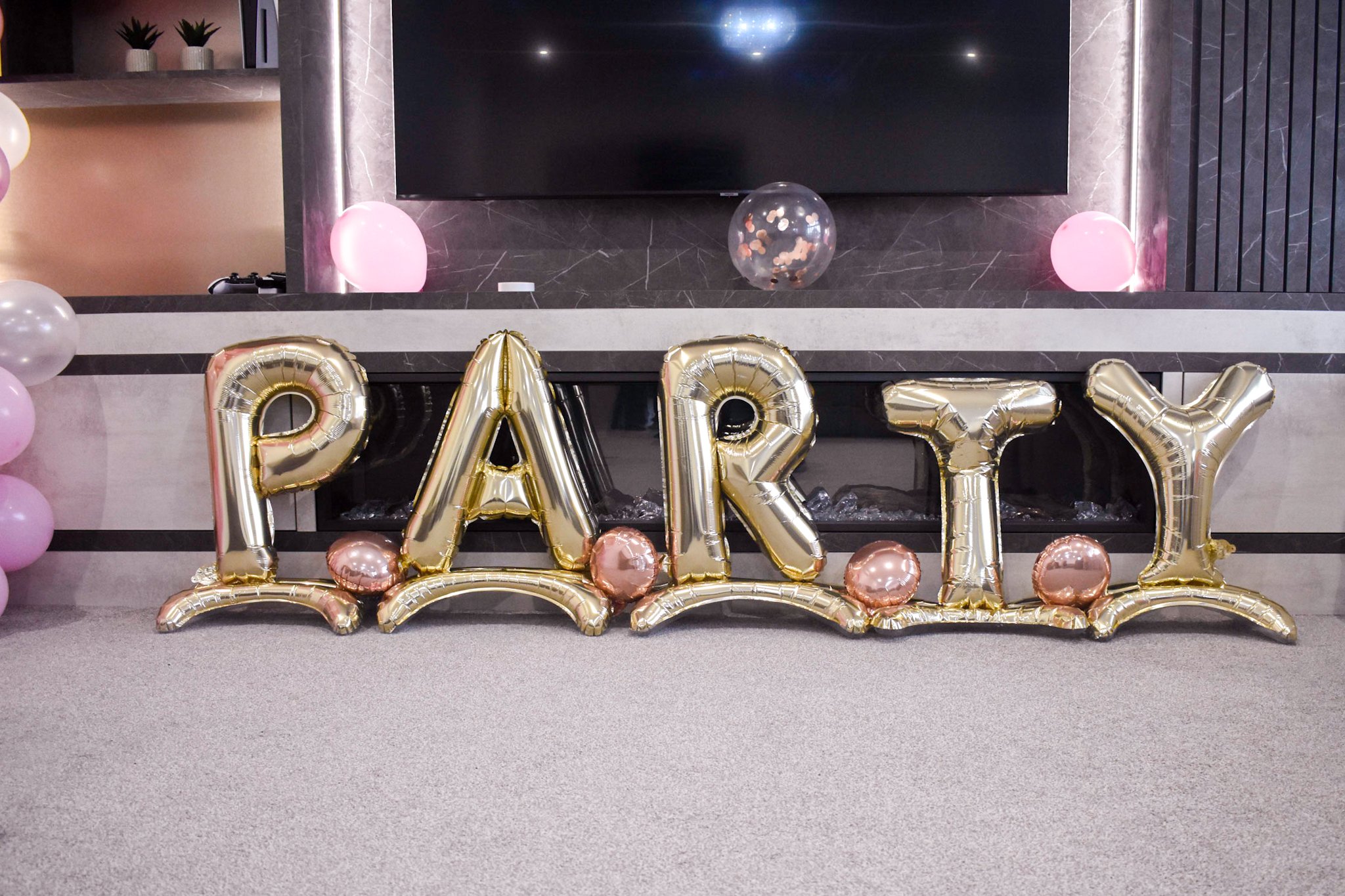 Golden balloon letters spell out 'PARTY' on a carpeted floor, surrounded by pink and rose gold balloons, with a modern fireplace and TV on the wall behind, in a decorated party setting.