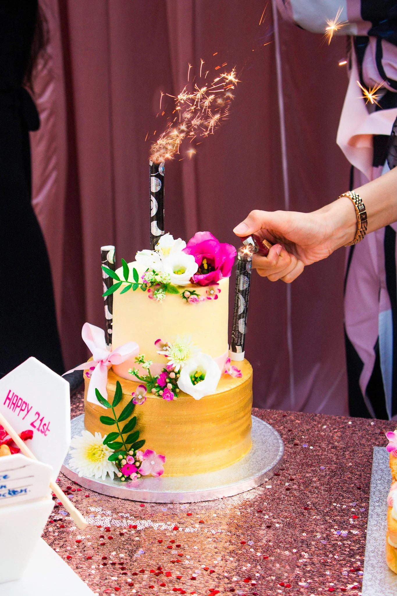 A person is lighting sparklers on a flower-decorated, two-tiered cake during a celebration, with a sign that reads 'HAPPY 21st' in the foreground.