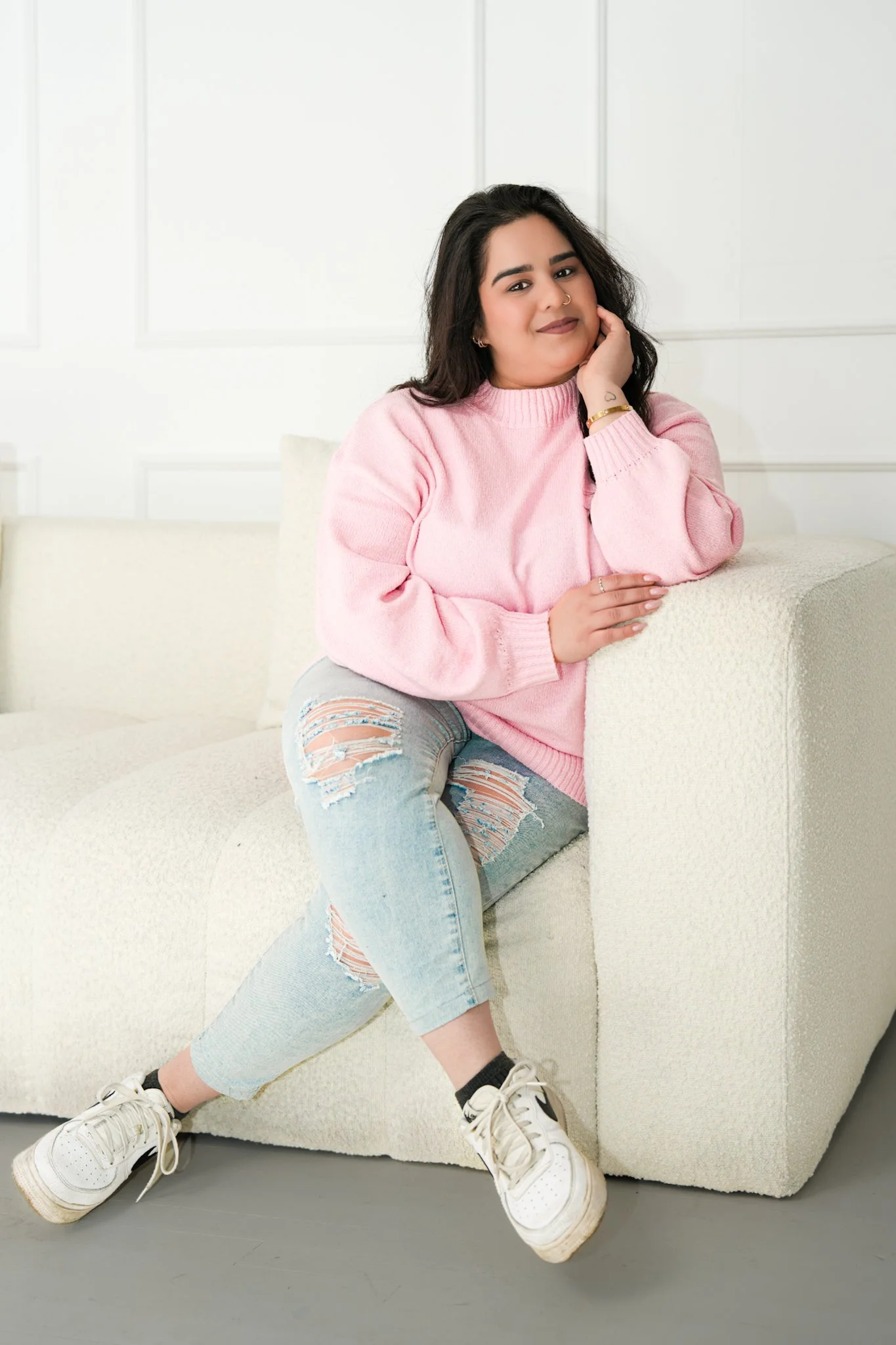A woman with dark hair wearing a pink sweater, ripped jeans, and sneakers sitting on a cream-colored sofa in a white room.
