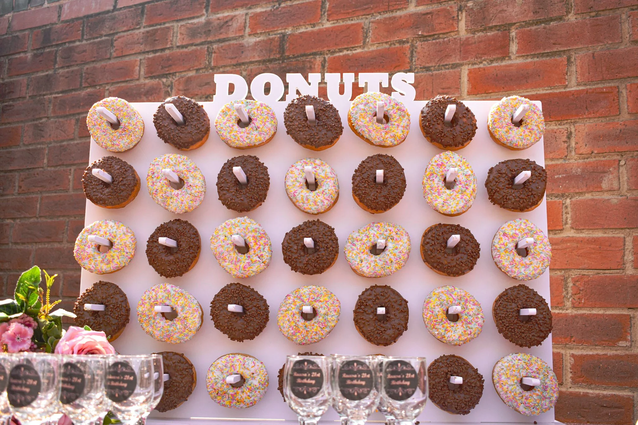 A display of assorted donuts, some with chocolate and some with rainbow sprinkles, arranged on a white board with a brick wall background. There is a sign that says 'DONUTS' at the top and some empty glasses with labels at the bottom.