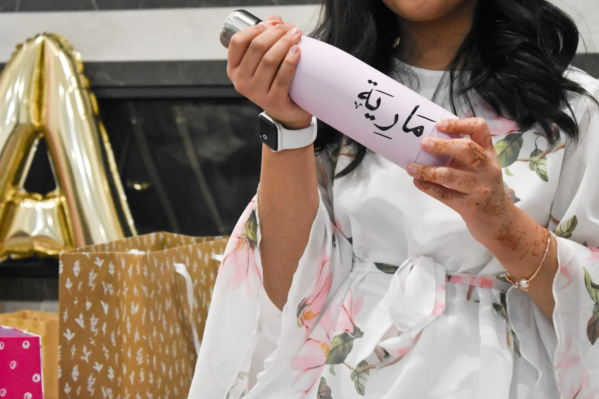 A woman with henna on her hands wearing a white floral robe holding a pink rolled-up paper with Arabic writing. There are gold letter balloons and gift bags on a table beside her.