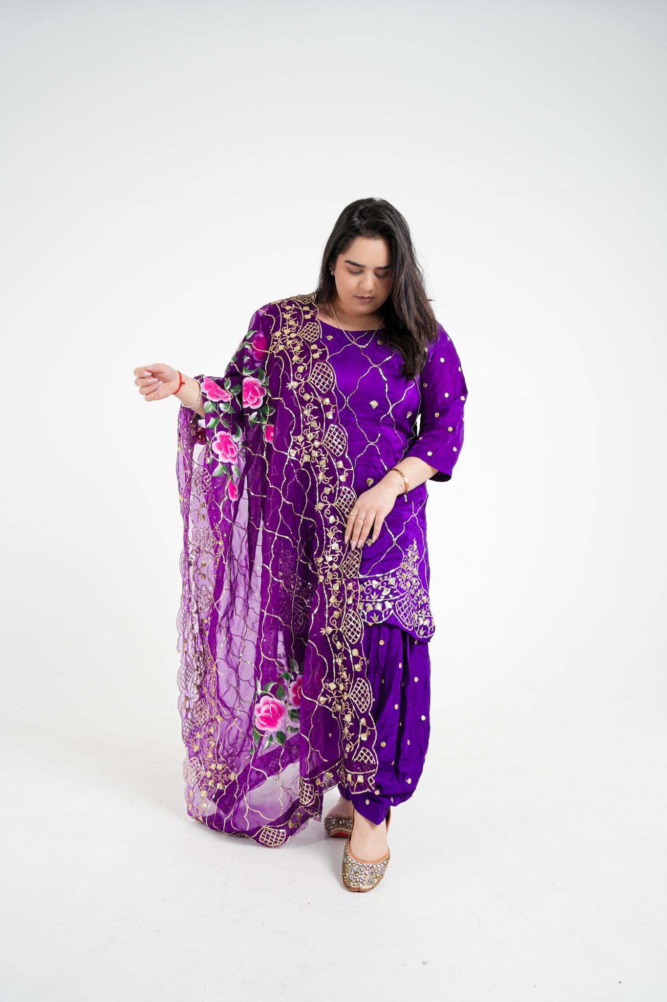A woman dressed in a purple traditional Indian outfit with gold embroidery and a sheer dupatta with floral embroidery, standing against a white background.