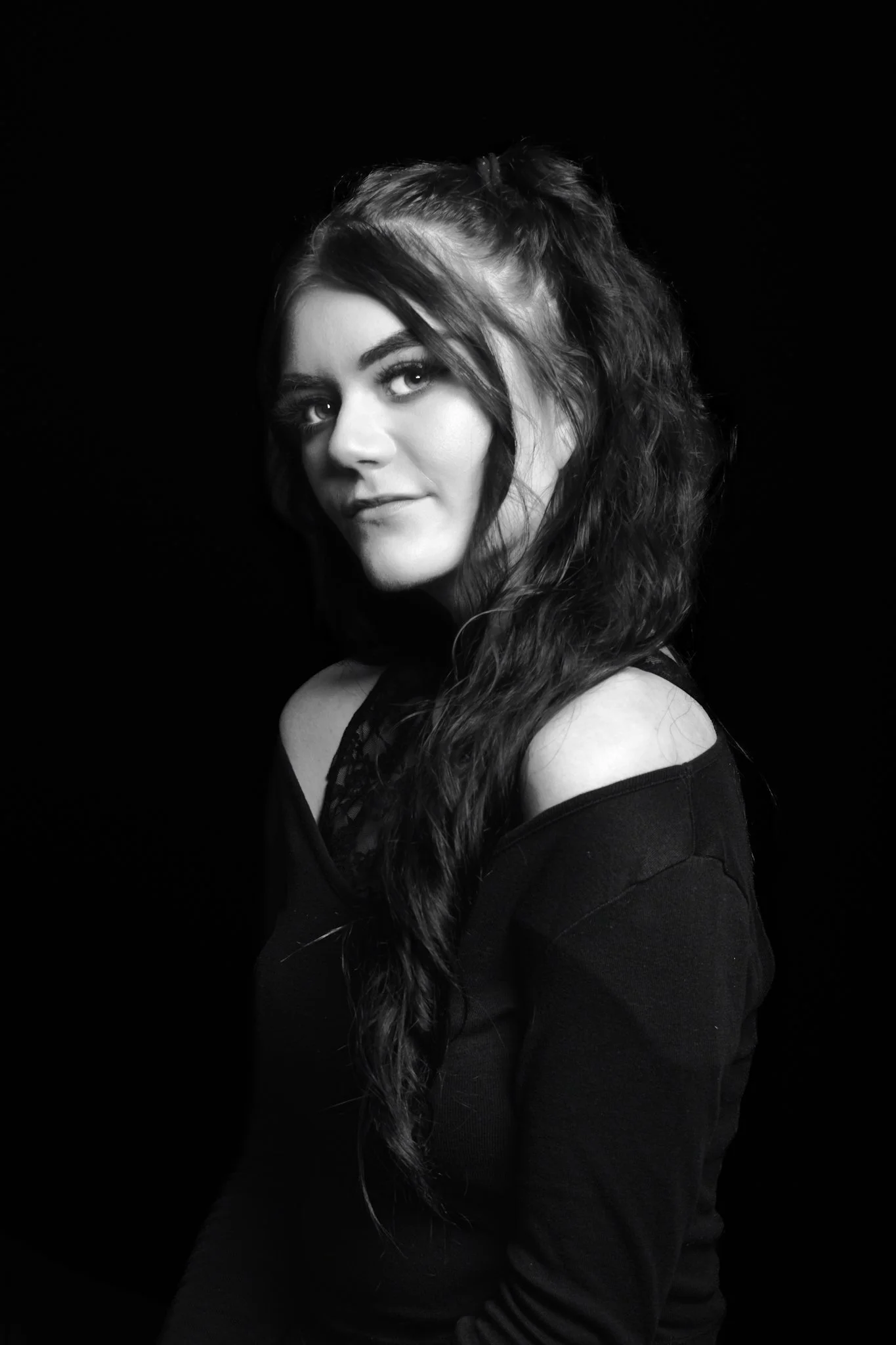 A black and white portrait of a young woman with long wavy hair, wearing a black top, against a dark background.