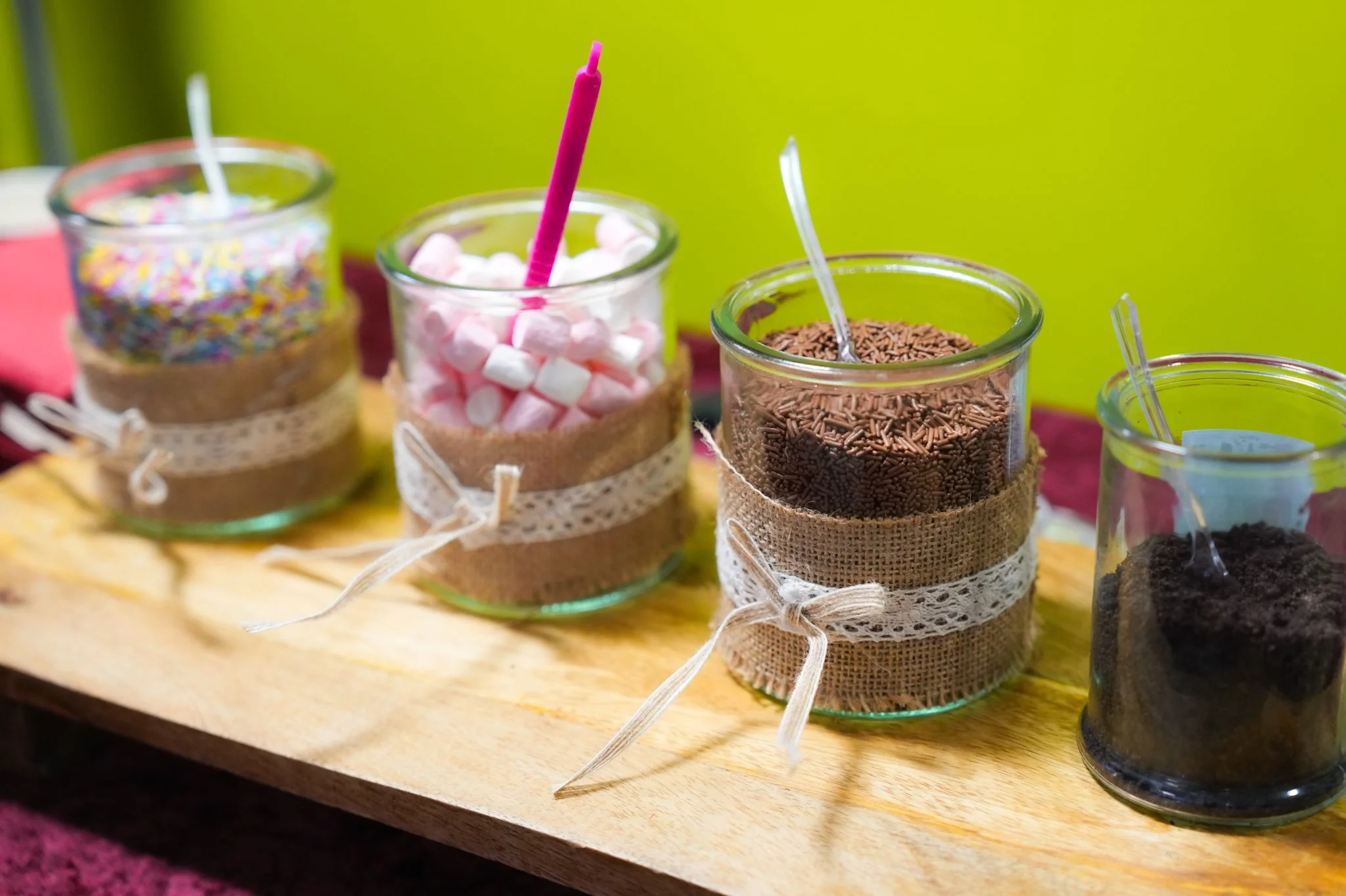Glass jars filled with various colored sprinkles, marshmallows, and chocolate sprinkles, decorated with burlap and lace ribbons, placed on a wooden surface.