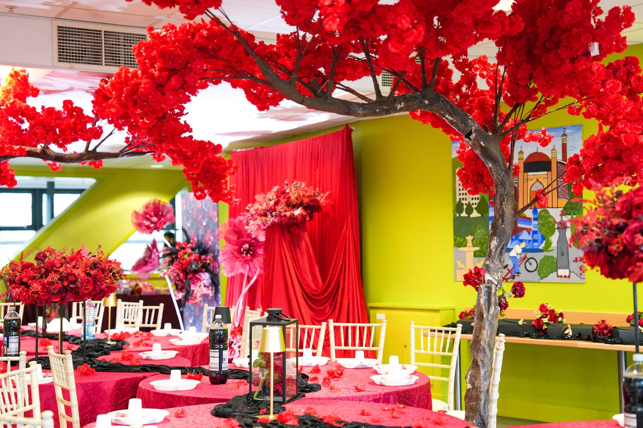 Interior decorated for celebration with red and black floral arrangements, red drapes, and a long table set with white plates, bottles, and candles. A red tree with artificial blossoms is in the foreground, with colorful wall art in the background.