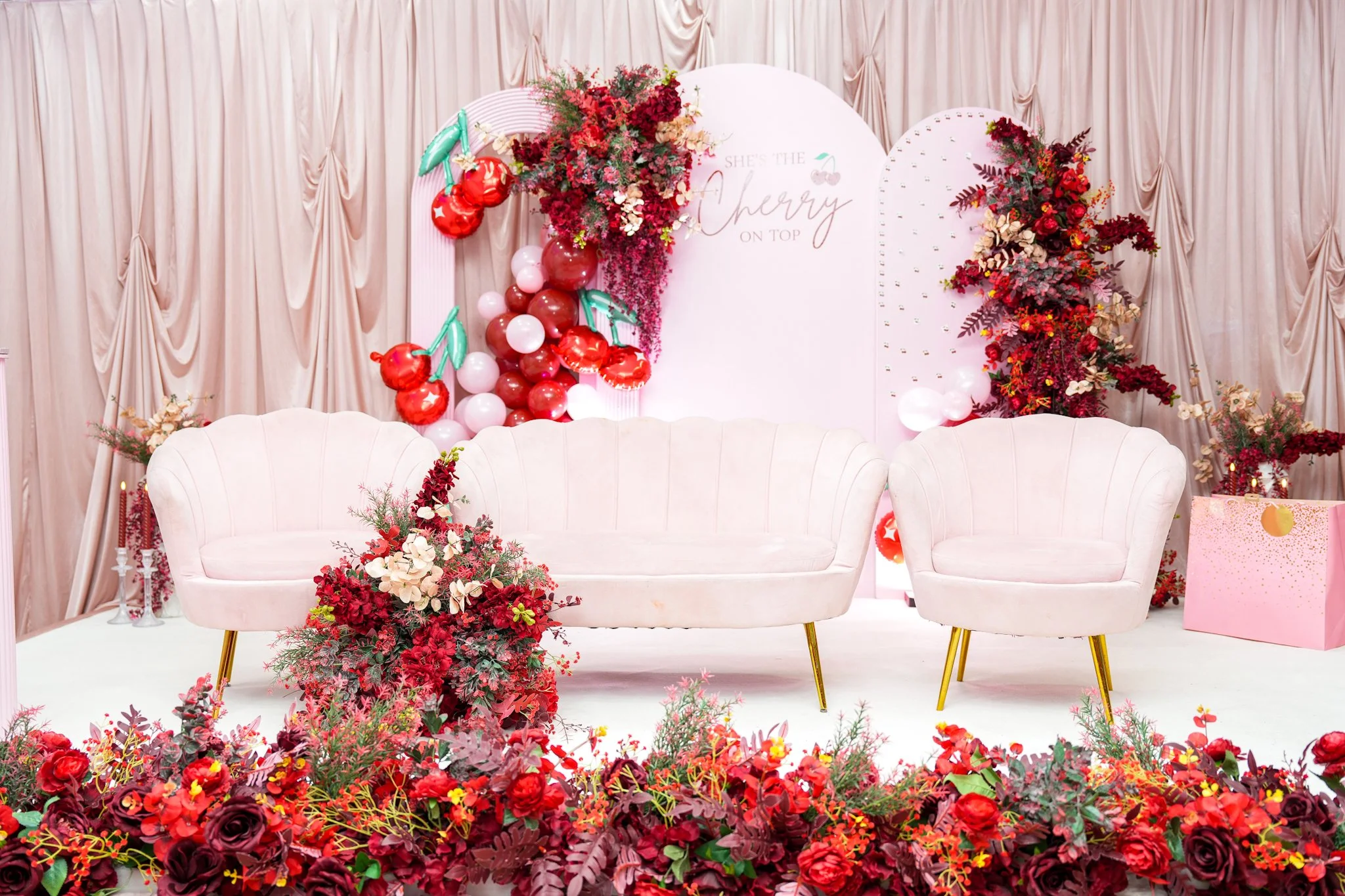 A decorated event space with pink velvet curtains, a white sofa, two pink armchairs, floral arrangements, balloons, and a backdrop with the text "She's the Cherry on Top."
